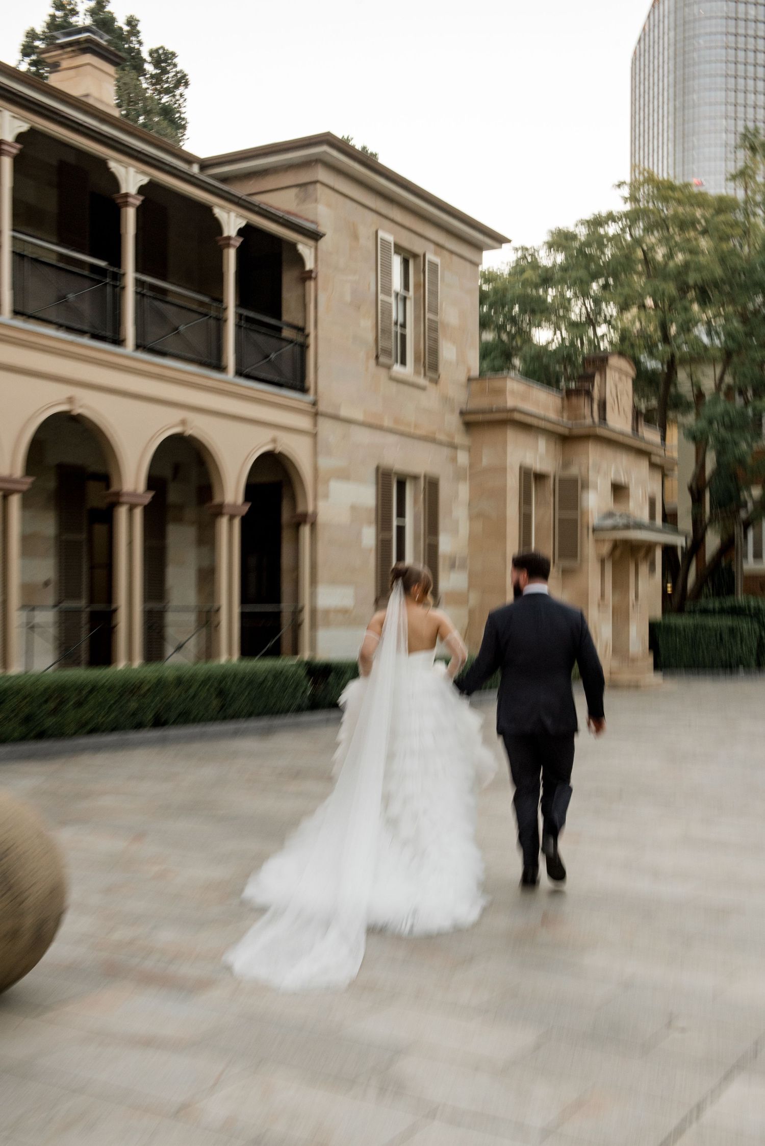 Bride and groom walking hand in hand past historic sandstone building at sunset.