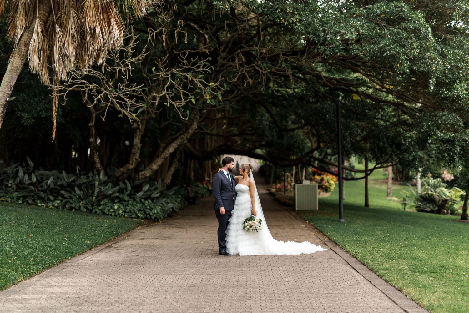 Bride and groom share a romantic kiss on a brick pathway beneath a canopy of trees in a park.