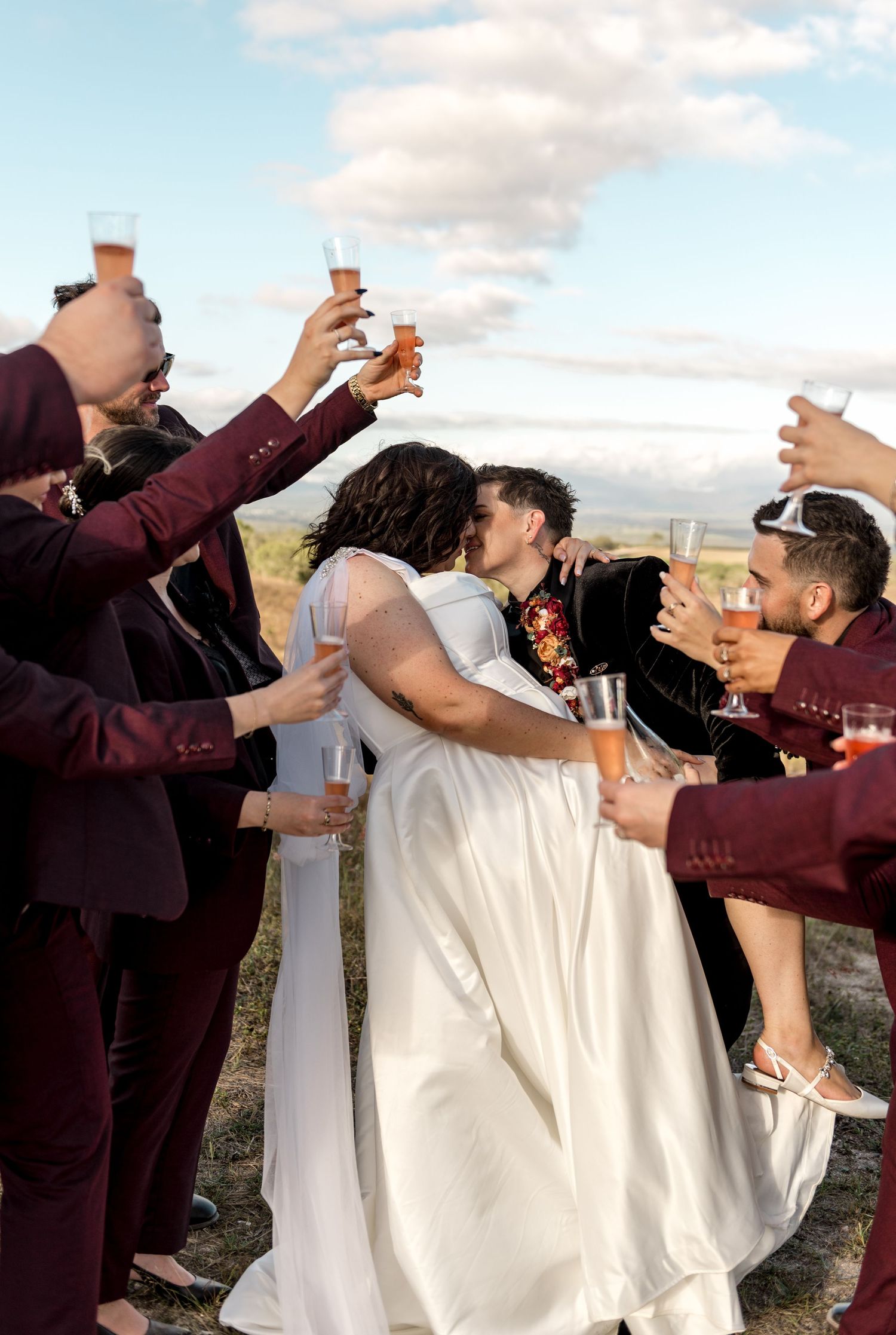 Wedding party toasting with champagne as bride and groom share a kiss outdoors.