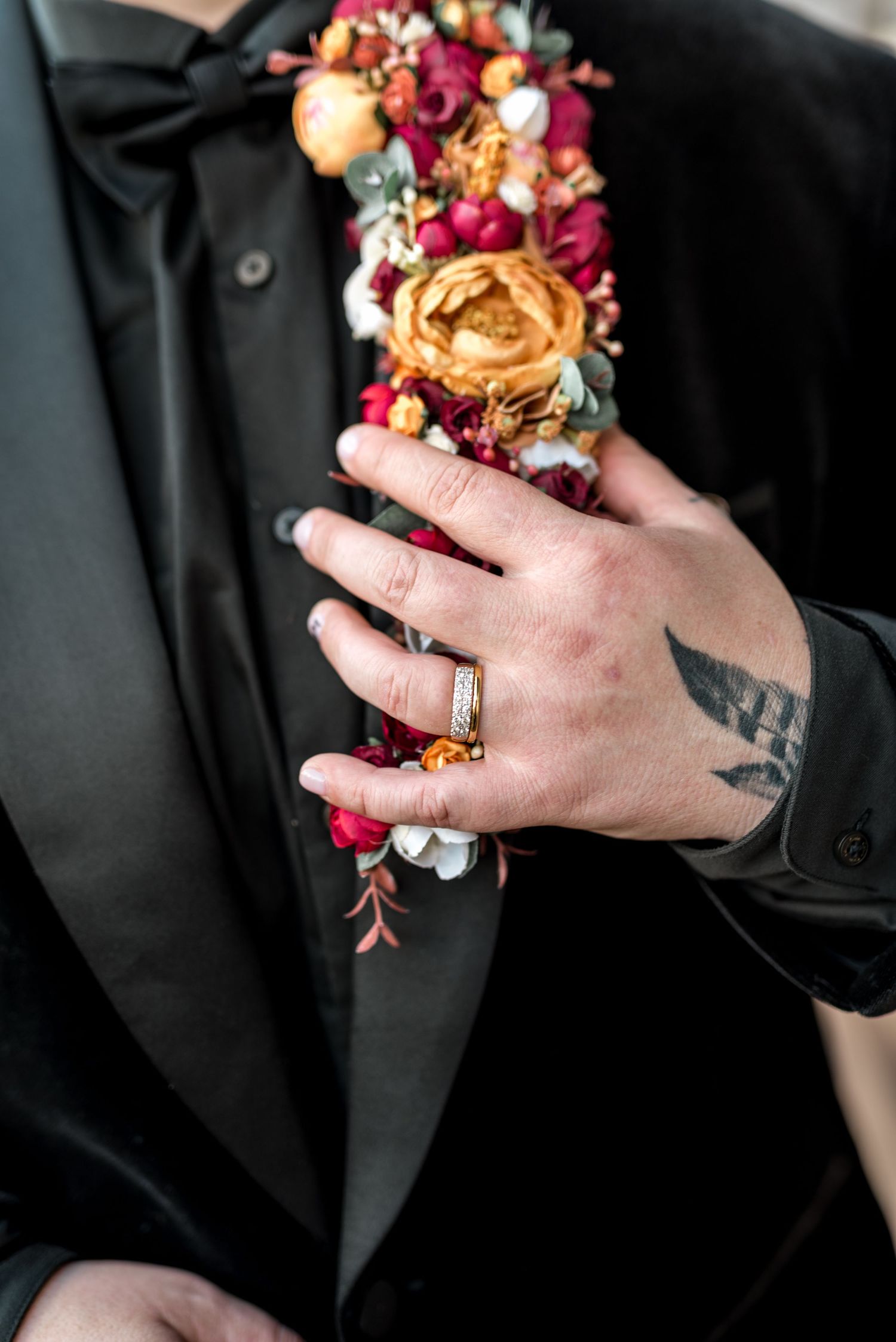 Close-up of tattooed hand with wedding ring touching colorful floral boutonniere on black tuxedo lapel.