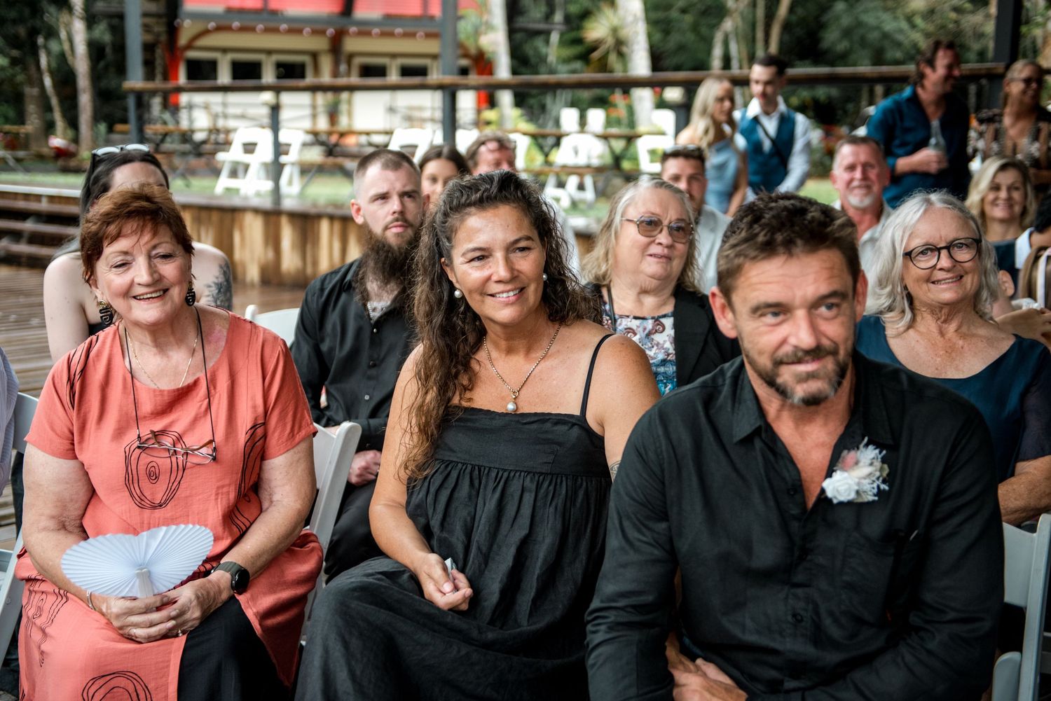 Wedding guests in black and coral attire seated and smiling at an outdoor ceremony.