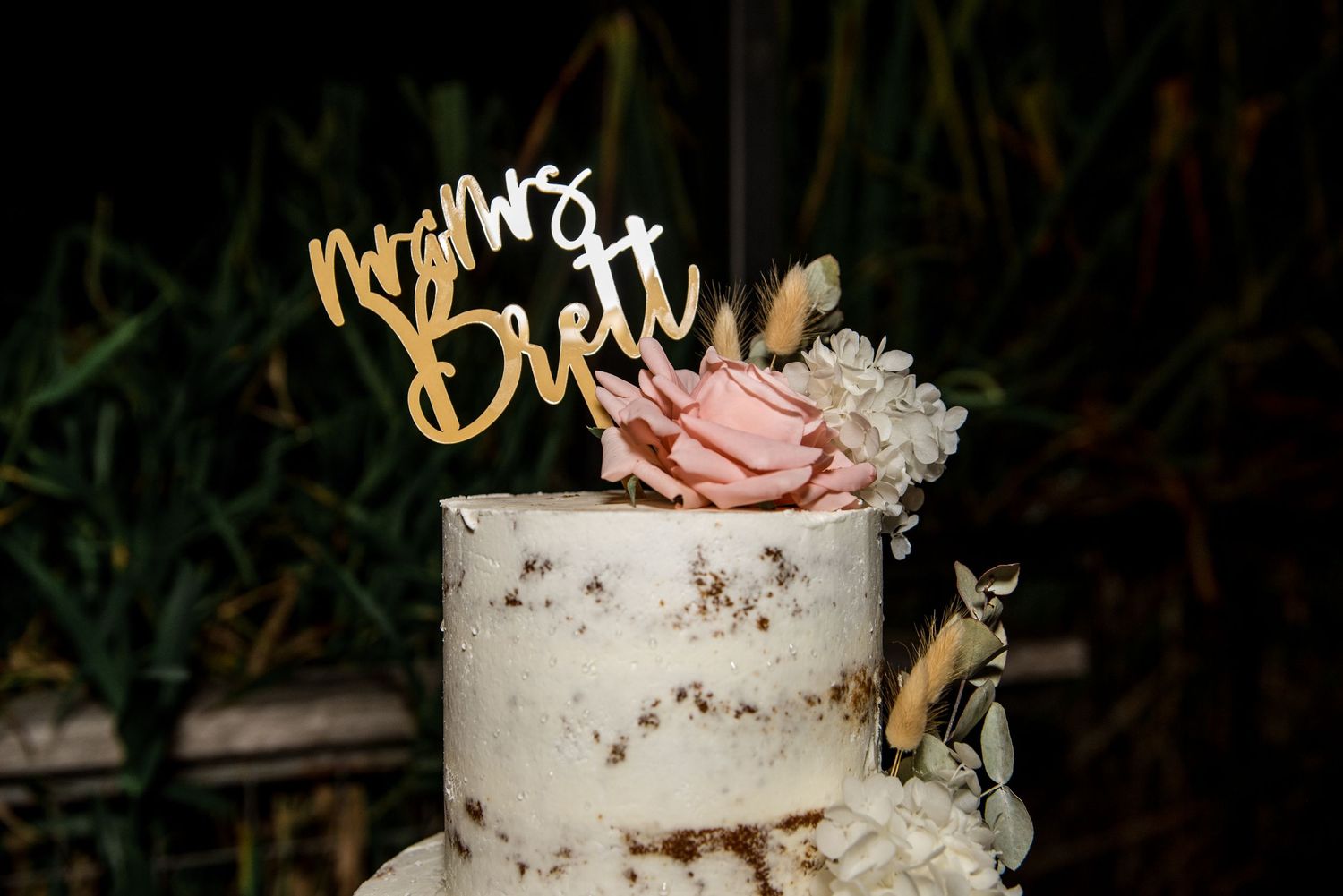 Two-tier white naked wedding cake topped with gold Mr and Mrs Brett topper and pink roses.