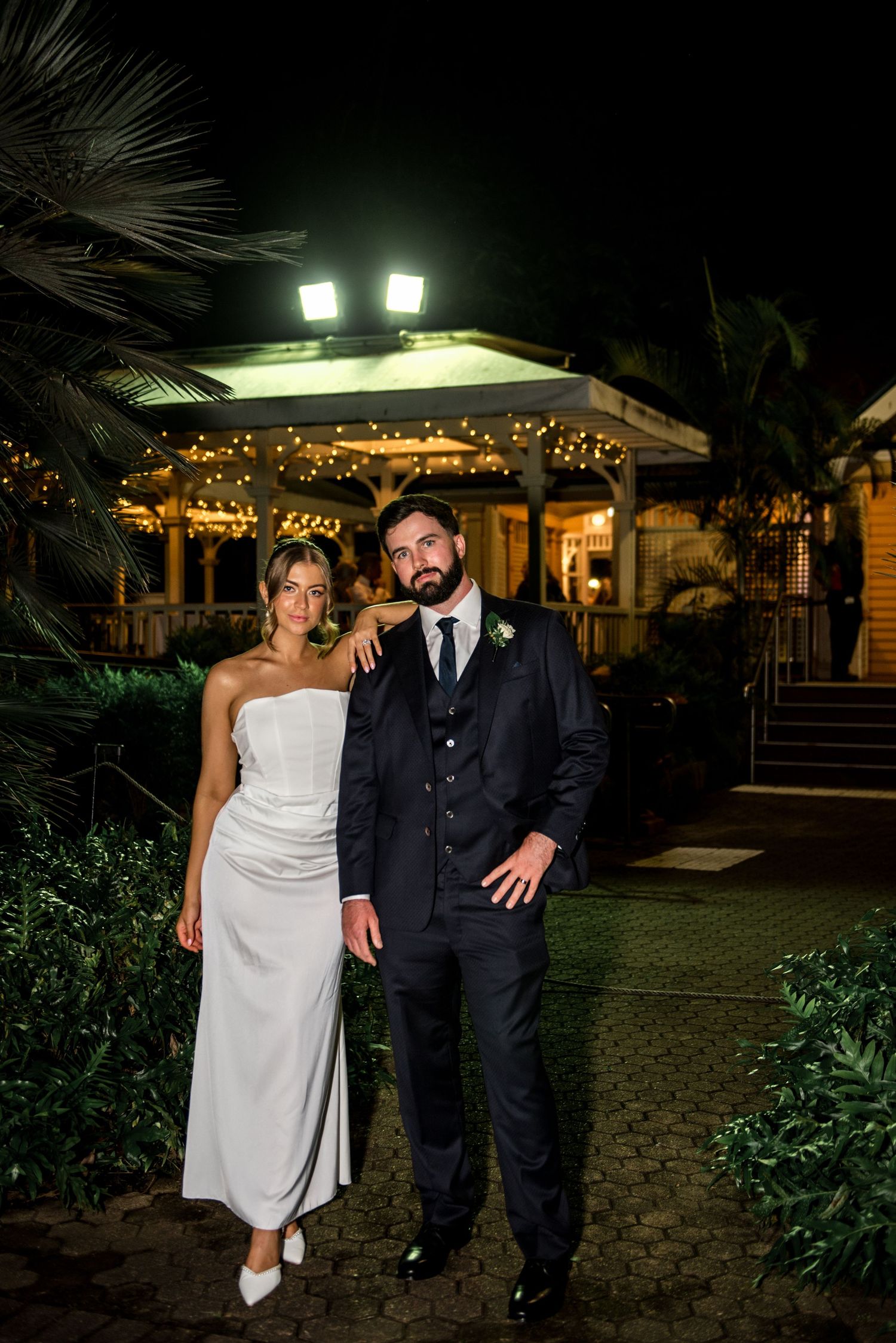Bride and groom posing together outdoors at night with festoon lights in the background.