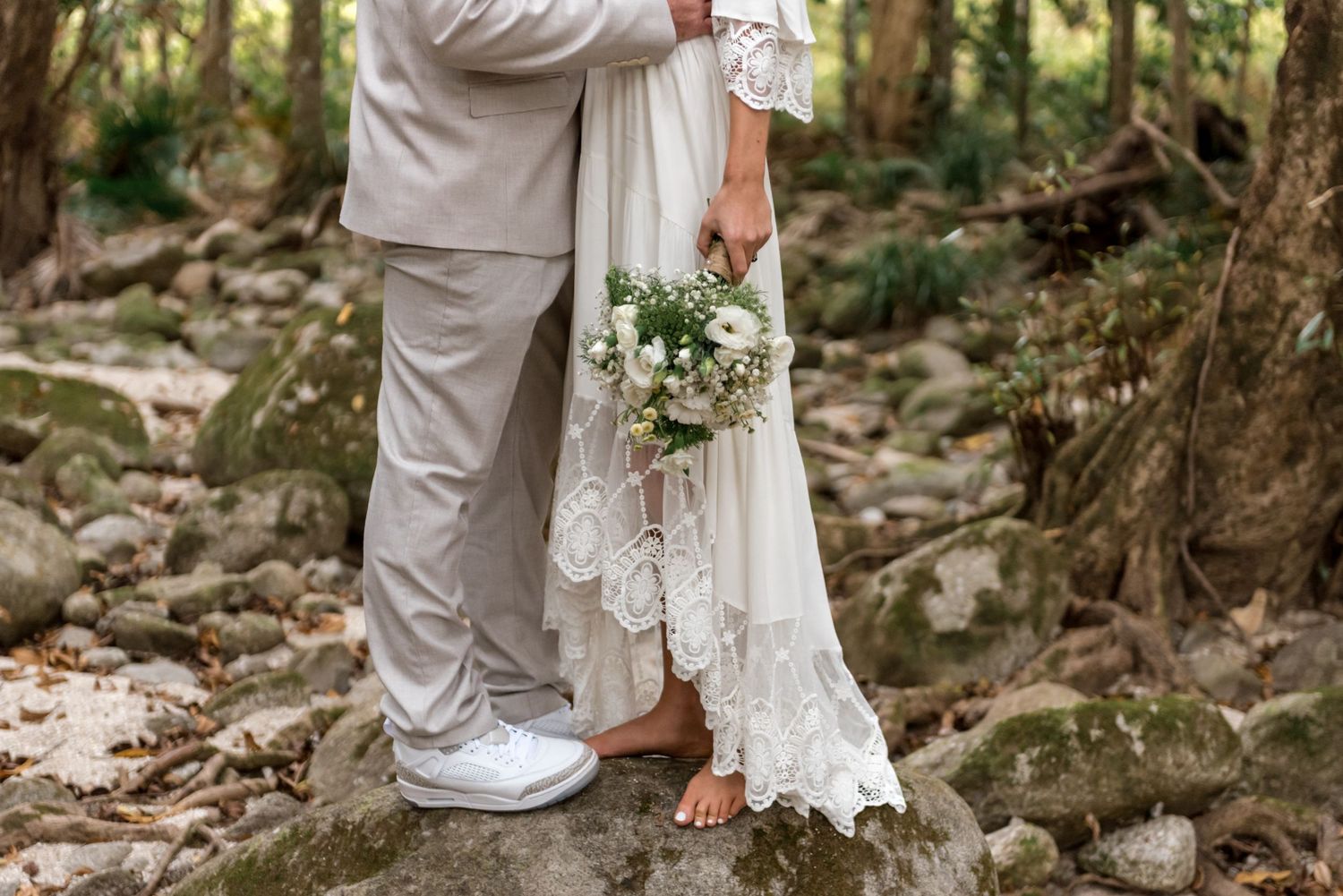 Bride and groom standing on rocks in forest, bride holding white bouquet, wearing lace wedding dress and groom in beige suit.