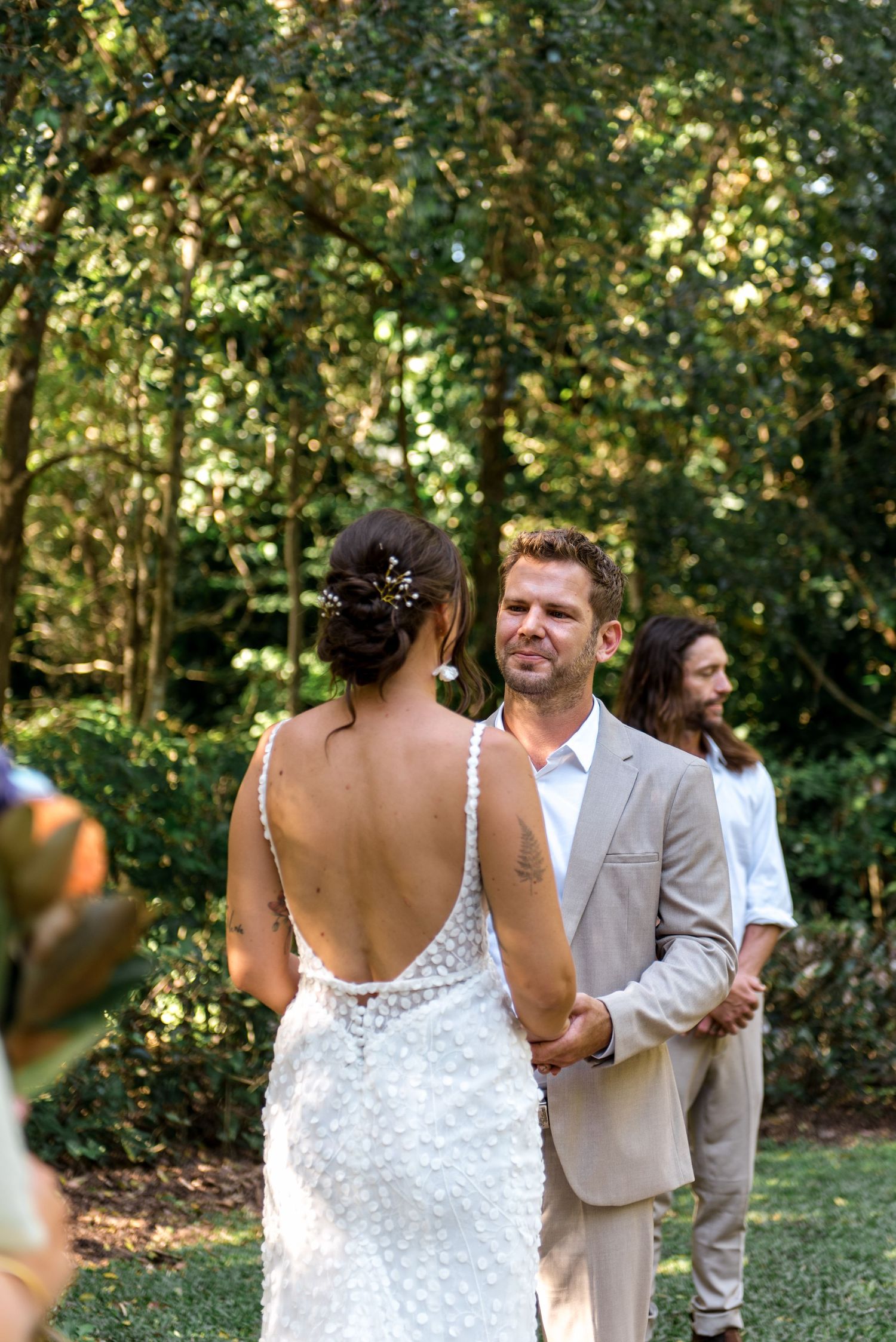 Groom in tan suit smiling at bride during outdoor garden wedding ceremony with officiant behind them.