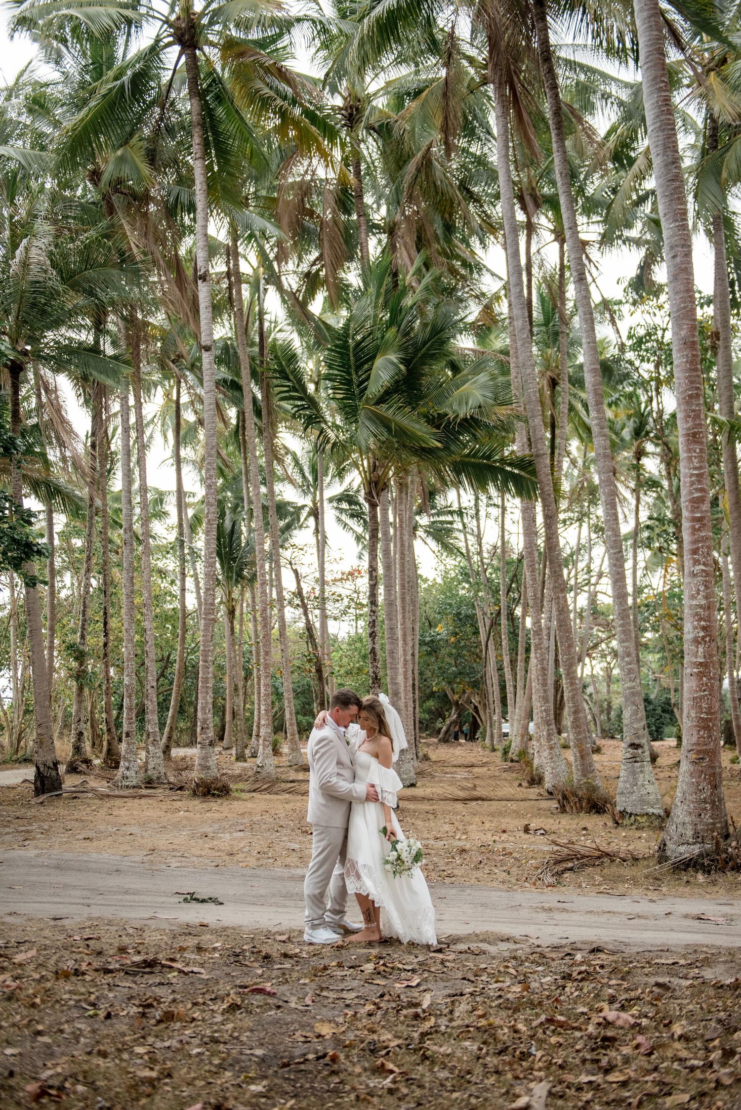 Bride and groom share a romantic kiss among tall coconut palm trees in tropical wedding setting.