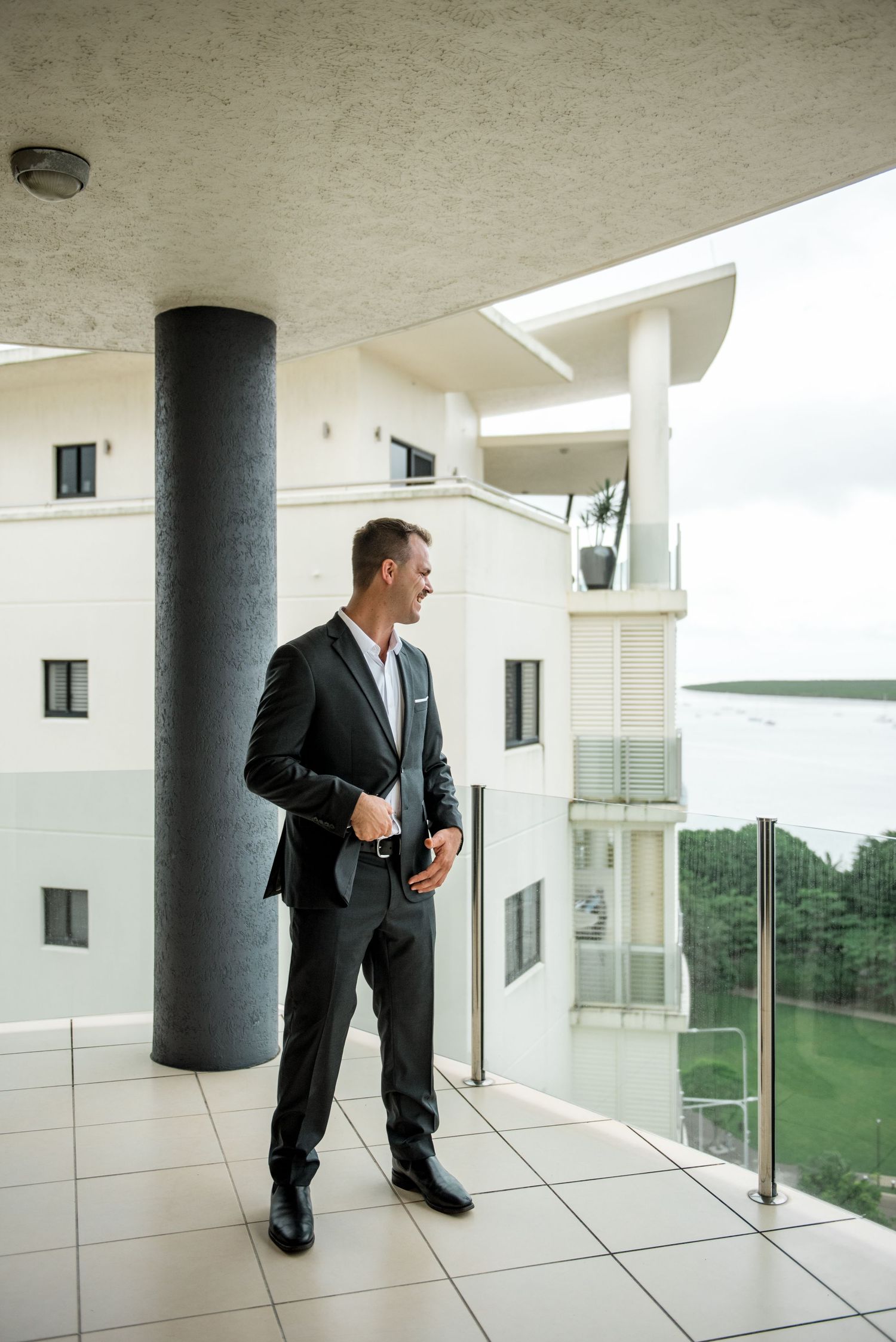 Dapper groom in dark suit stands confidently on a high-rise balcony overlooking a waterfront cityscape.