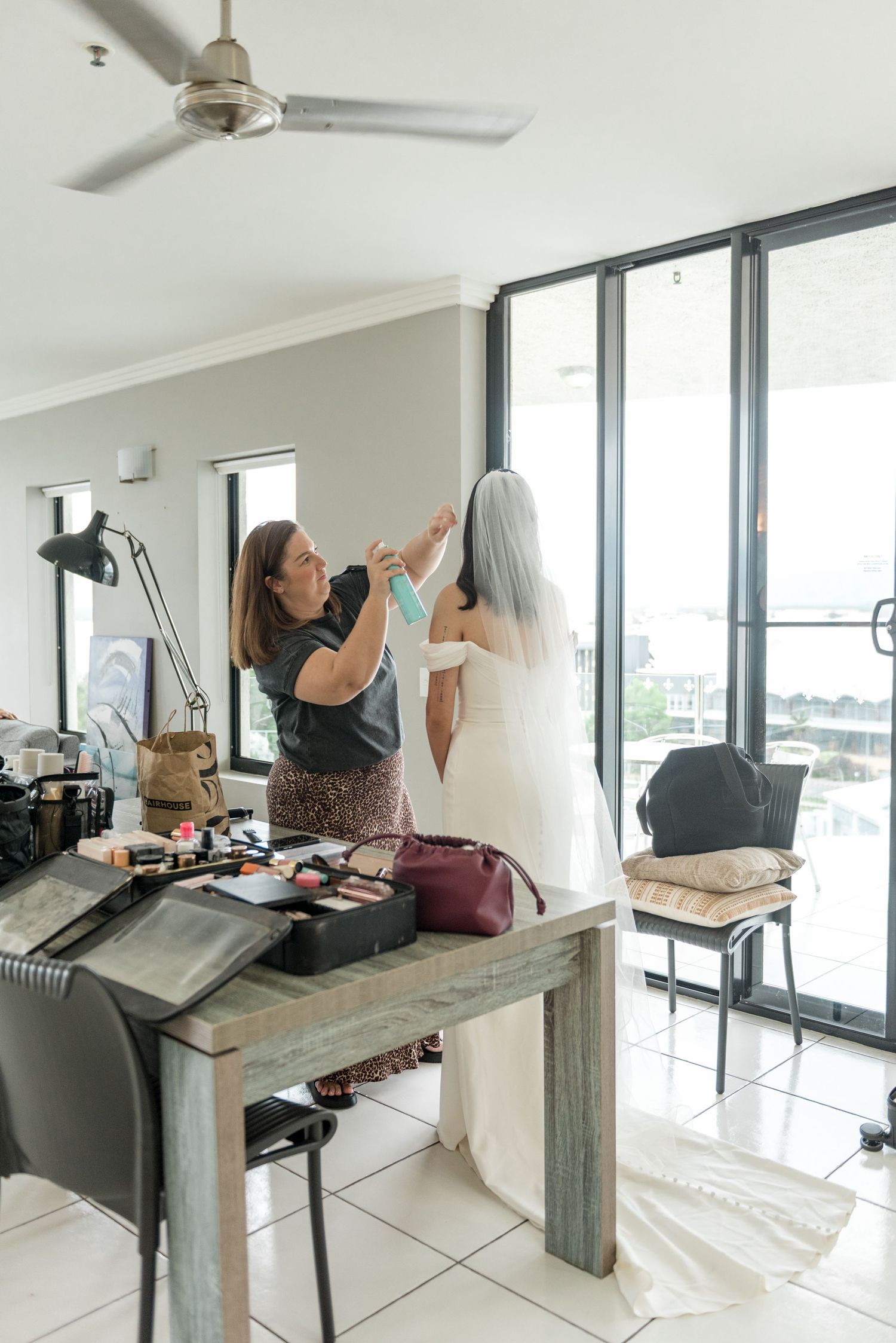 Bridal assistant helping bride with veil in bright room with makeup table nearby.