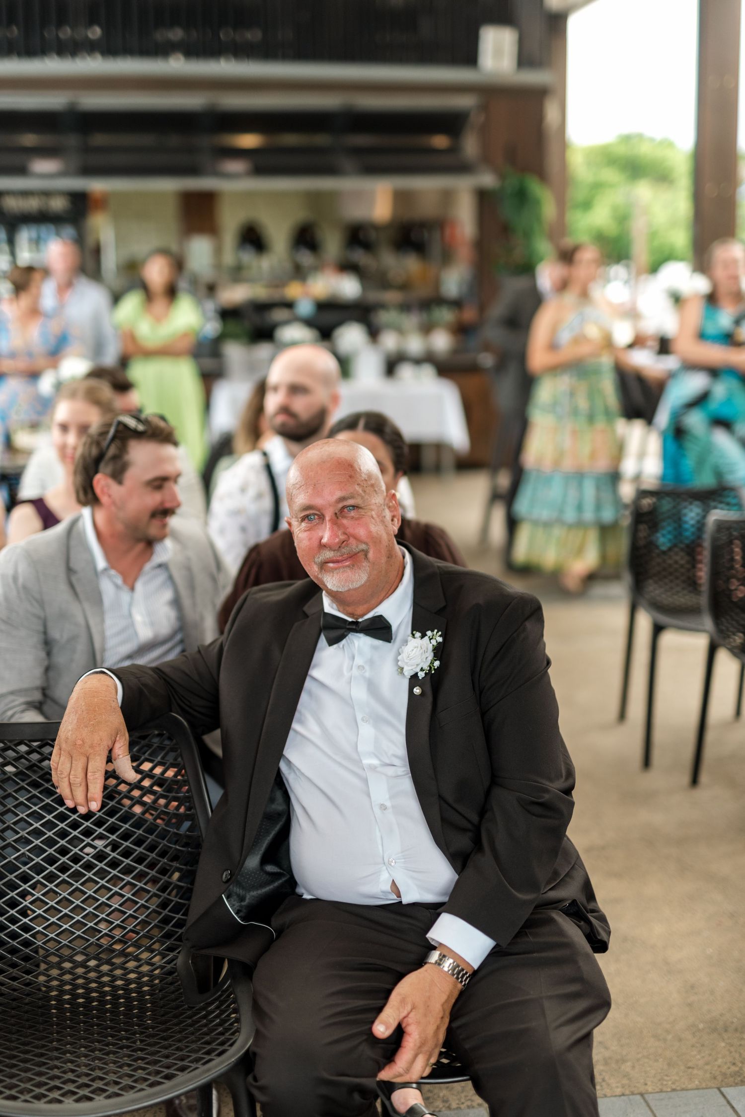 Elderly man in tuxedo with bow tie and boutonniere smiling at wedding reception outdoors.