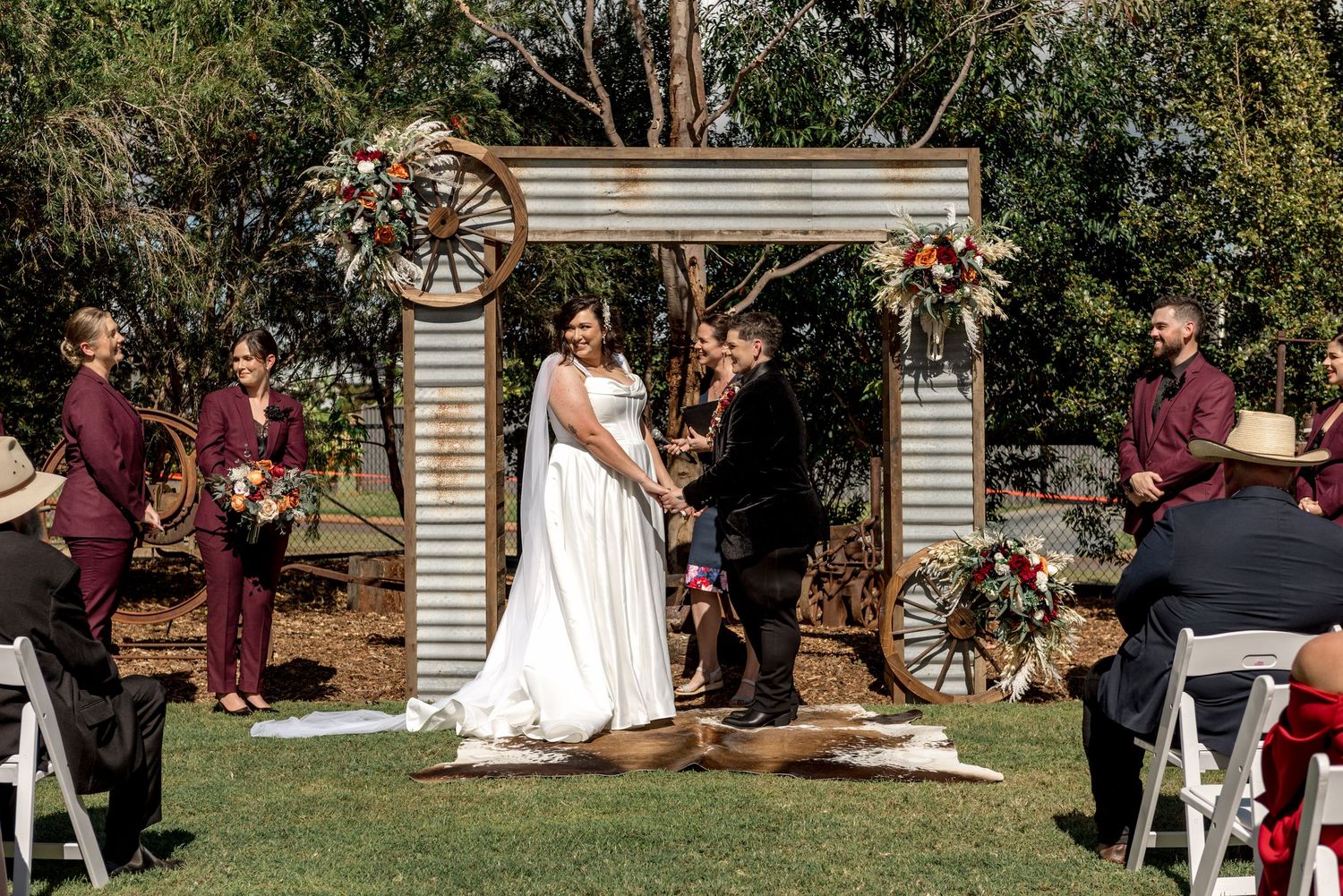 Bride and groom hold hands at rustic corrugated iron altar decorated with wagon wheels and colorful floral arrangements.