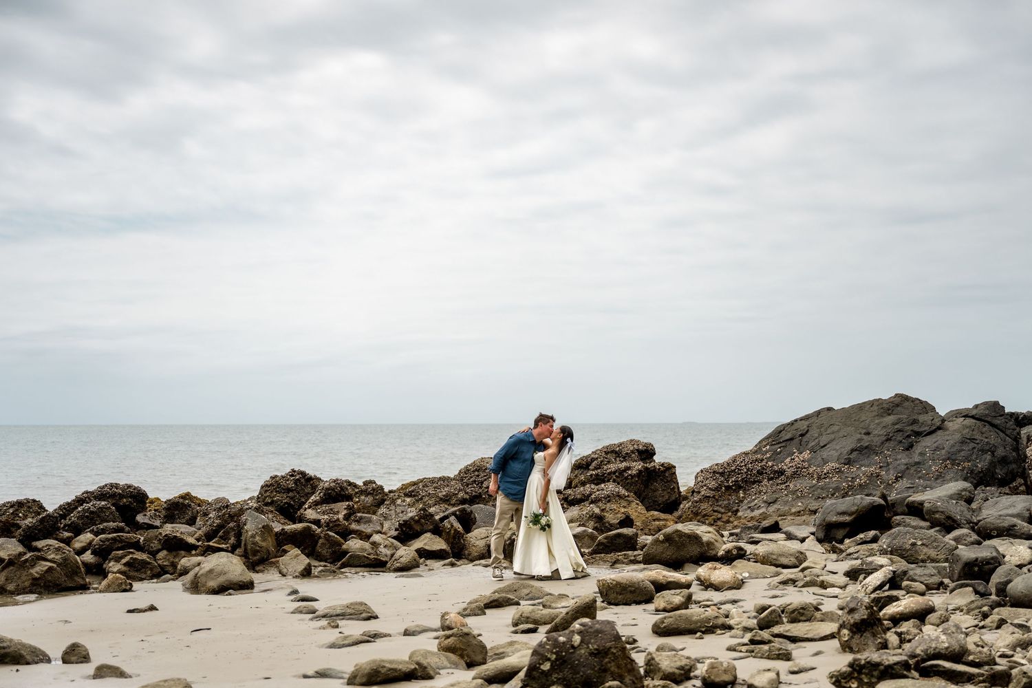 Bride and groom share romantic kiss on rocky beach shoreline under overcast sky at Little Cove, Port Douglas.
