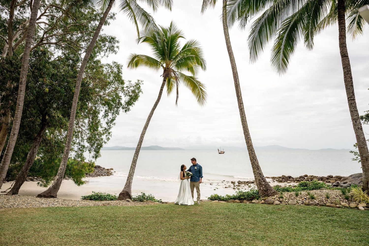 Couple stands beneath palm trees on a tropical beach wedding venue with ocean views at Little Cove, Port Douglas.