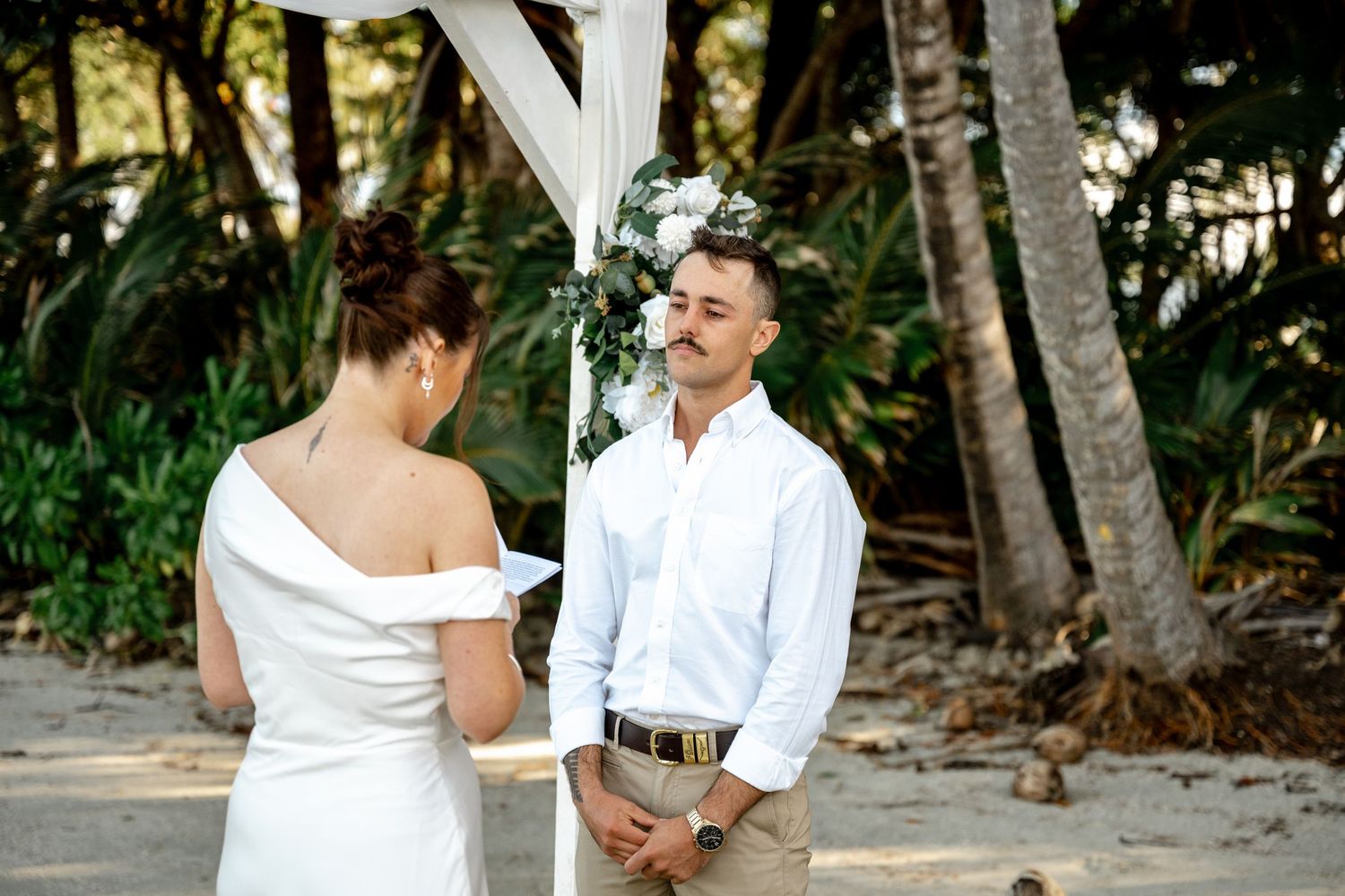 Groom listening attentively as bride reads vows at a tropical beach wedding ceremony under a floral arch at Four Mile Beach, Port Douglas.