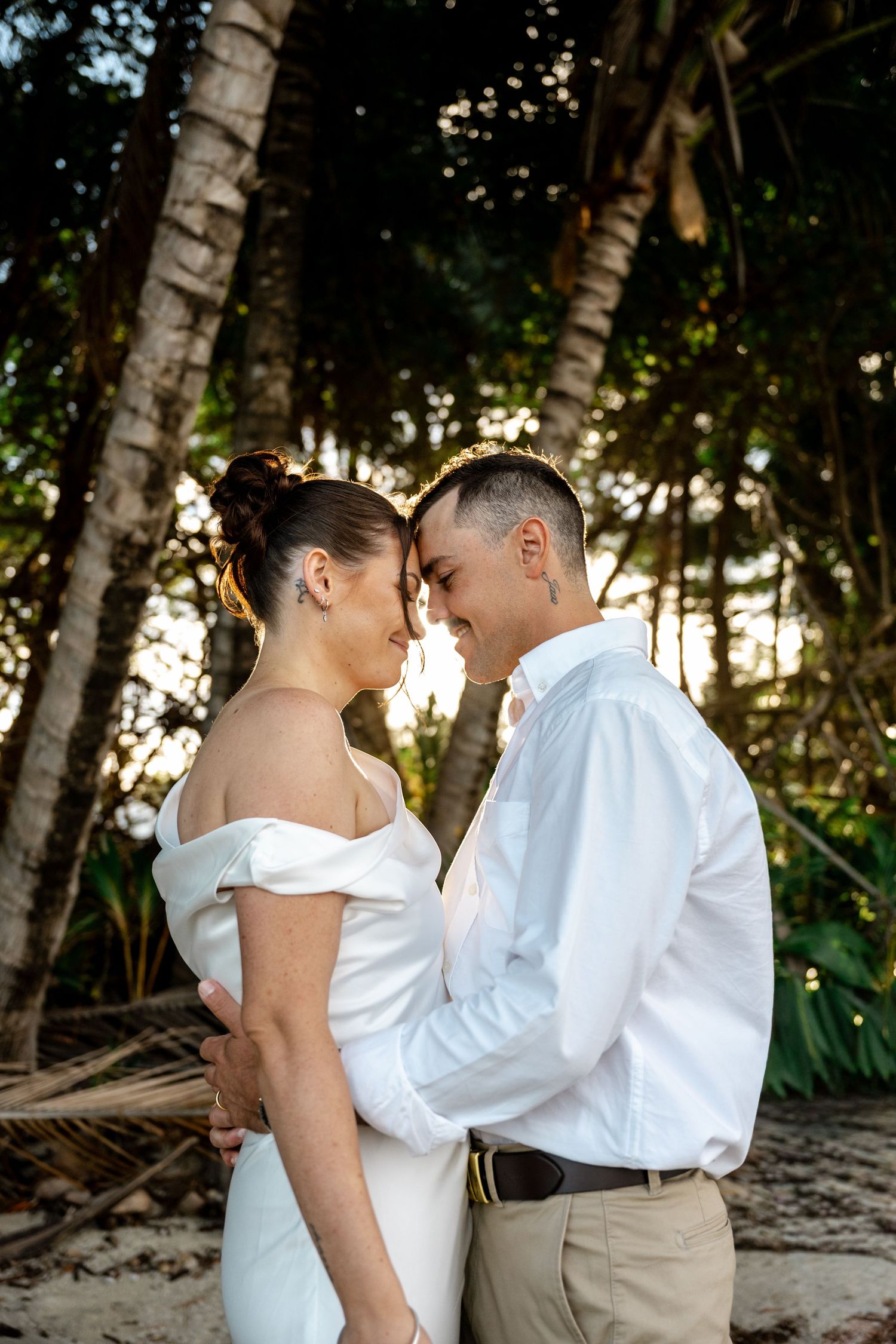 Bride and groom touch foreheads tenderly while embracing on a sandy beach among tropical palms at Four Mile Beach, Port Douglas.