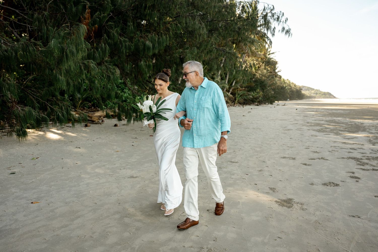Bride and groom walking together on sandy beach, surrounded by tropical greenery, bride carrying white floral bouquet at Four Mile Beach.
