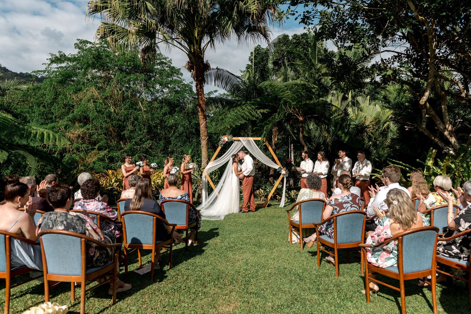 Outdoor wedding ceremony with guests seated on wooden chairs facing a couple at a floral arch.
