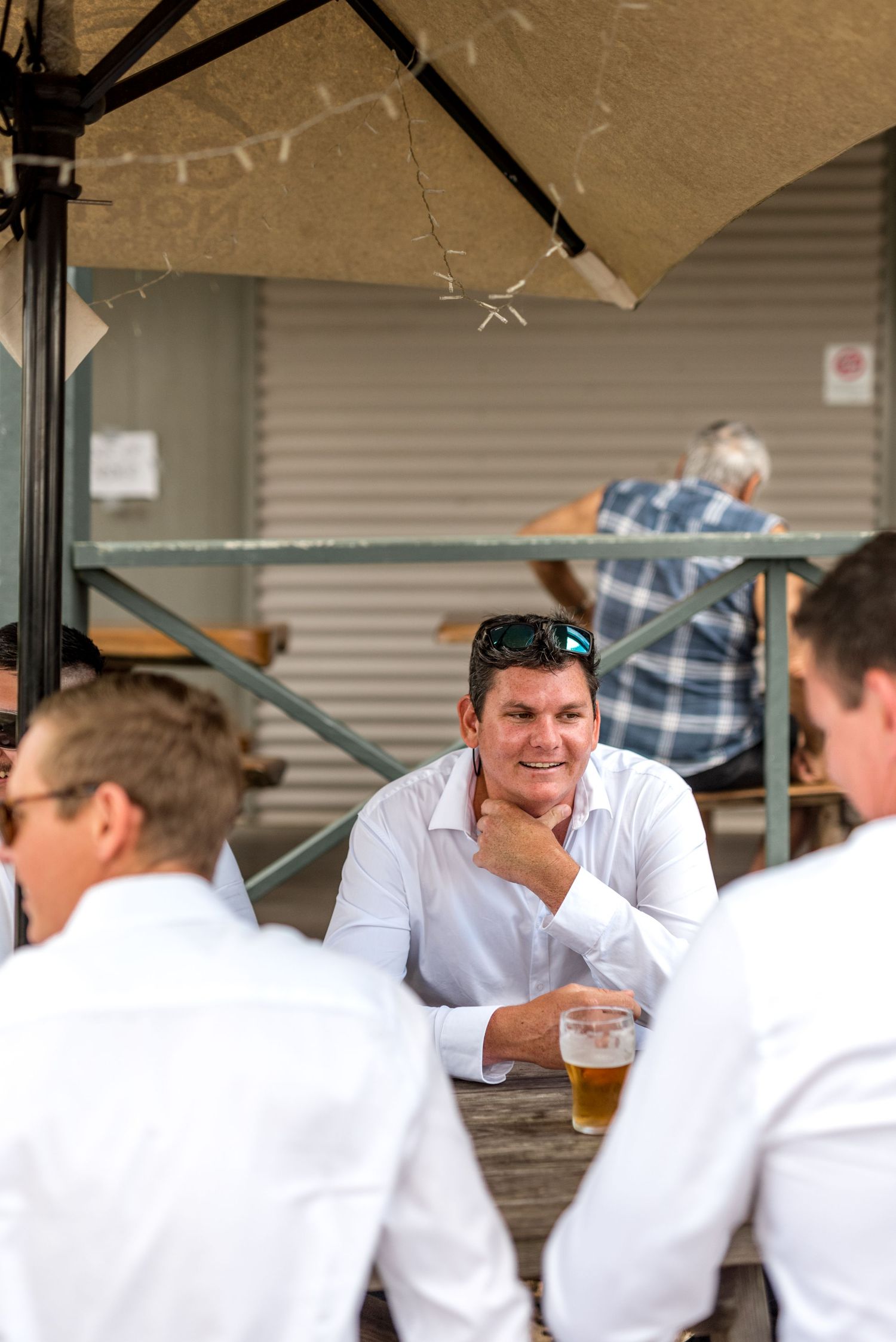Smiling groomsman in white shirt with sunglasses on head holding a beer at an outdoor wedding gathering.
