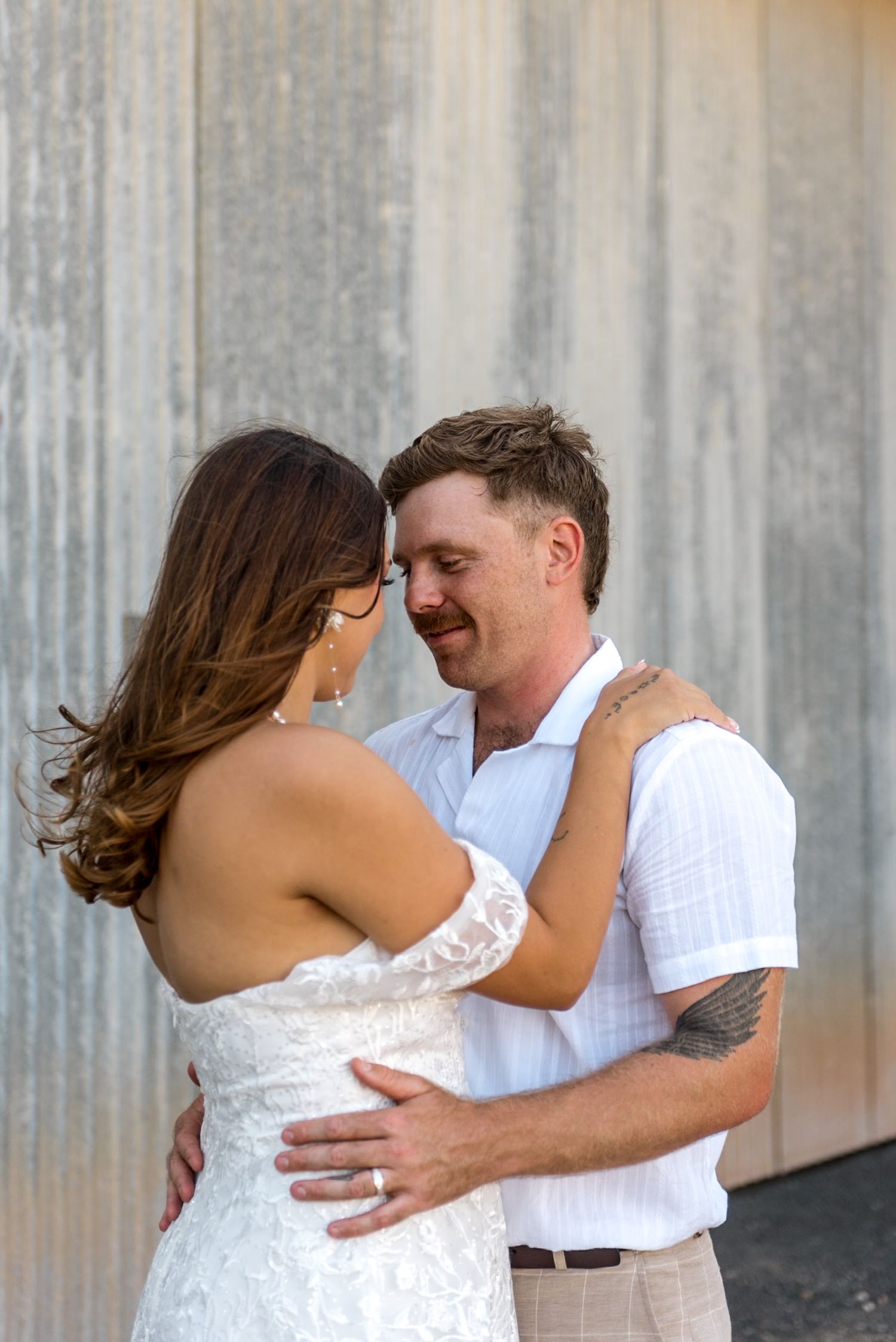 Smiling groom in white shirt tenderly holds bride in off-shoulder lace wedding gown against metal wall.