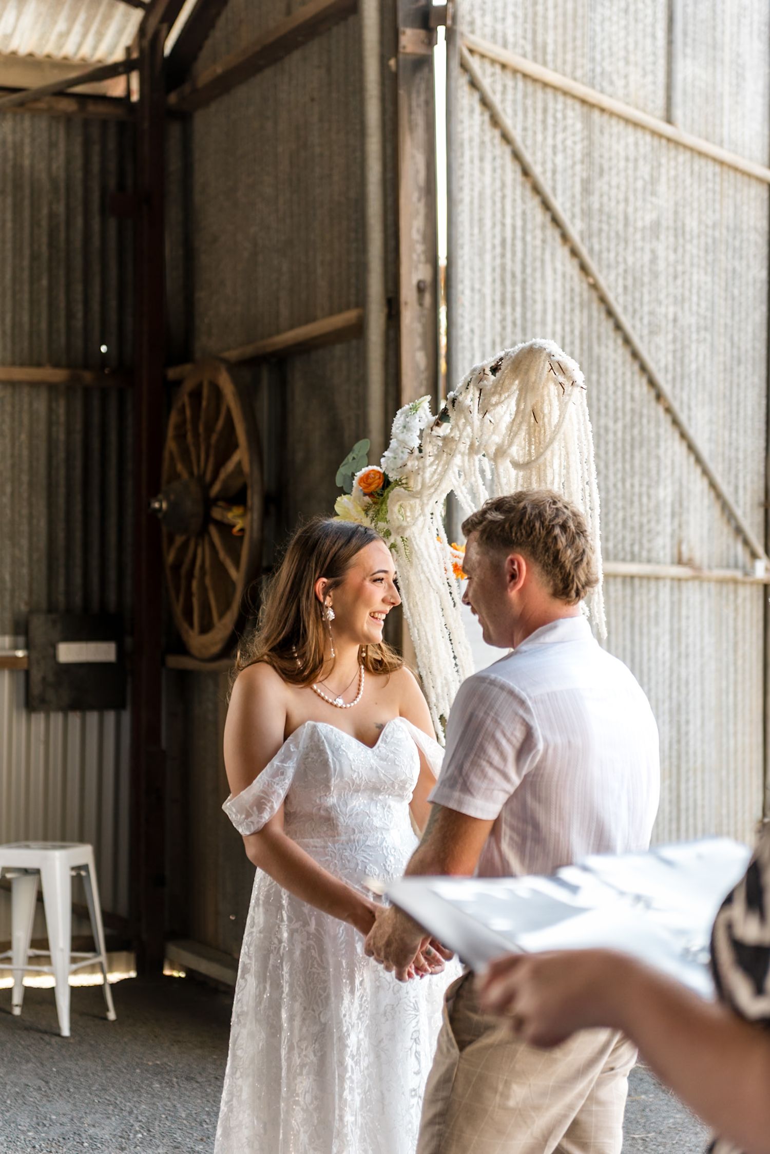 Bride laughing joyfully while holding grooms hands during heartfelt barn wedding ceremony beneath macrame arch.
