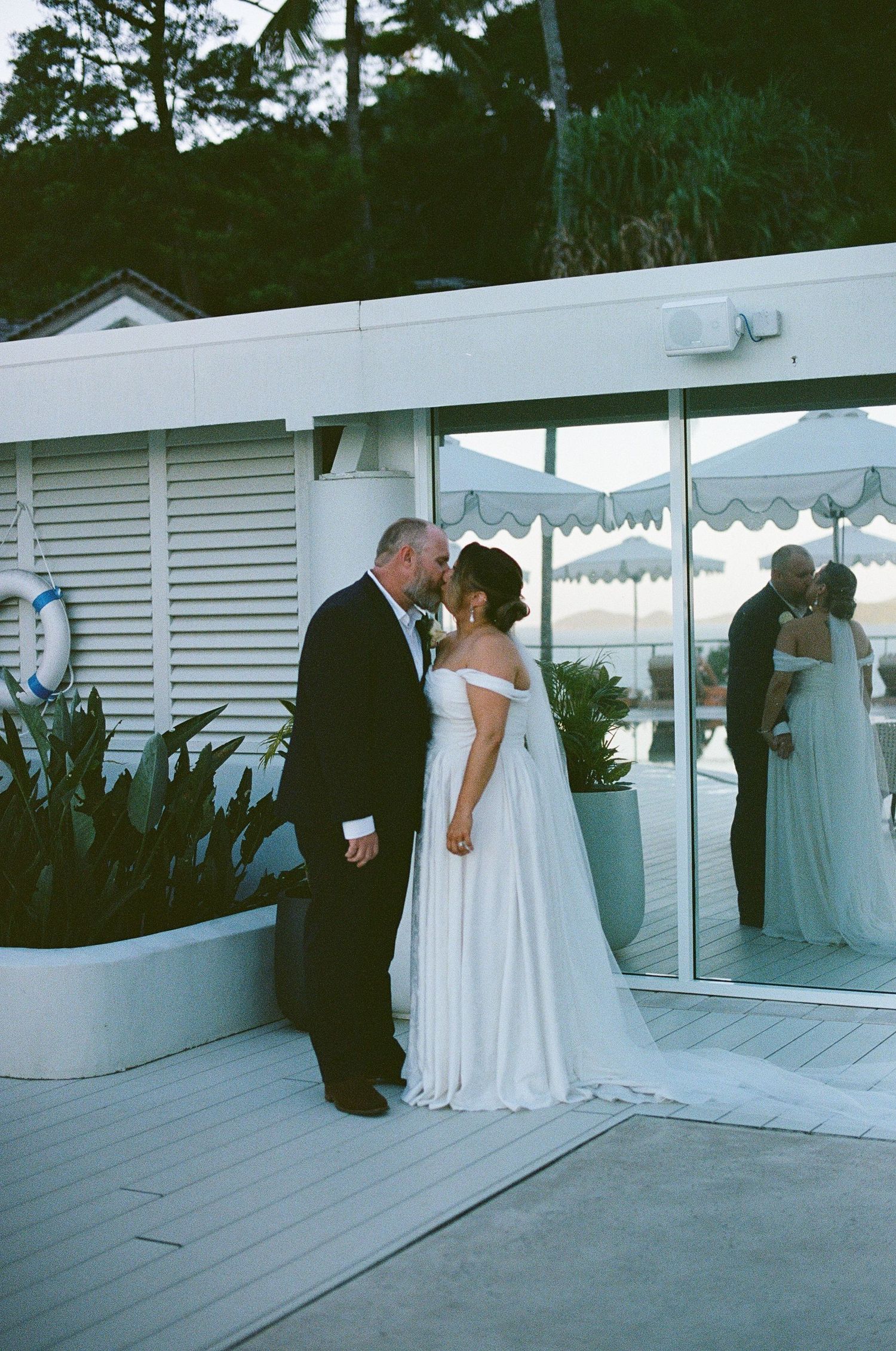 Bride and groom sharing a kiss outside a white beachside building at dusk.