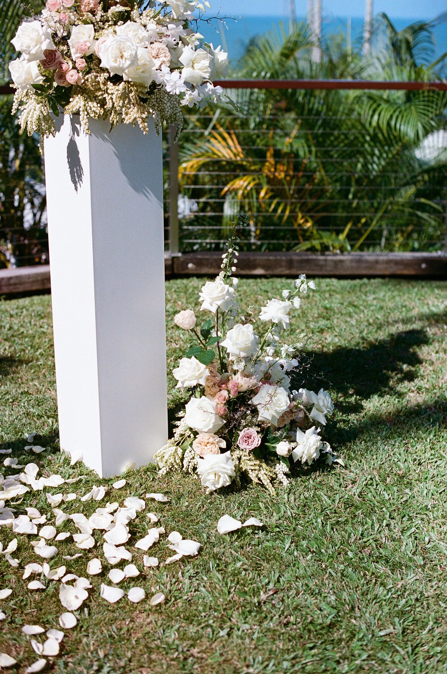 White wedding ceremony pillar adorned with blush and white floral arrangements on green lawn with rose petals.