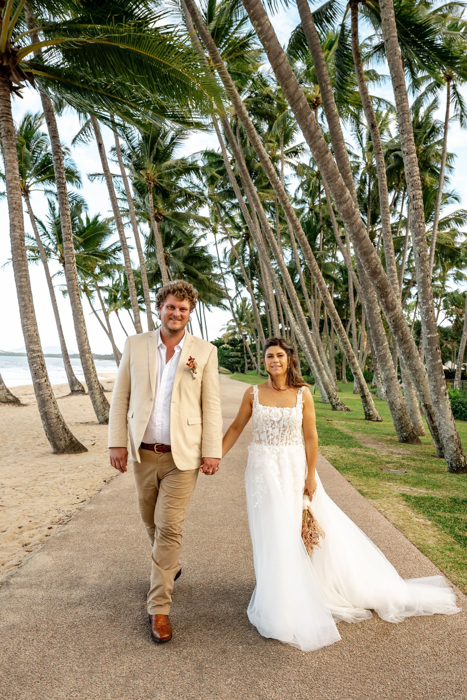 Bride in white gown and groom in tan suit walk hand-in-hand along a palm tree-lined beach path.