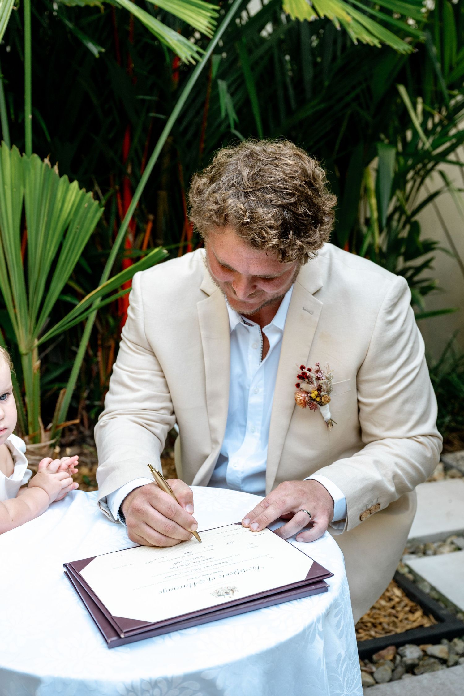 Groom in beige suit signing wedding certificate at outdoor table surrounded by tropical greenery.