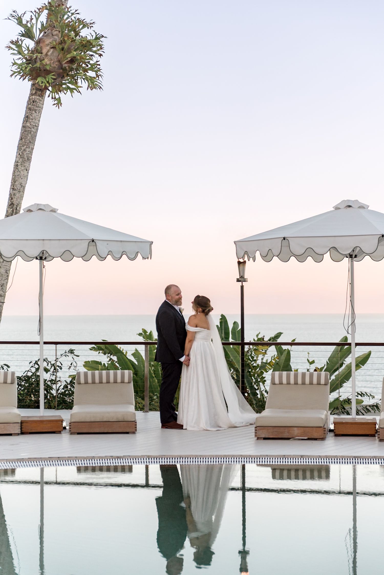Wedding couple kissing poolside at sunset with white scalloped umbrellas and ocean backdrop reflecting in water.