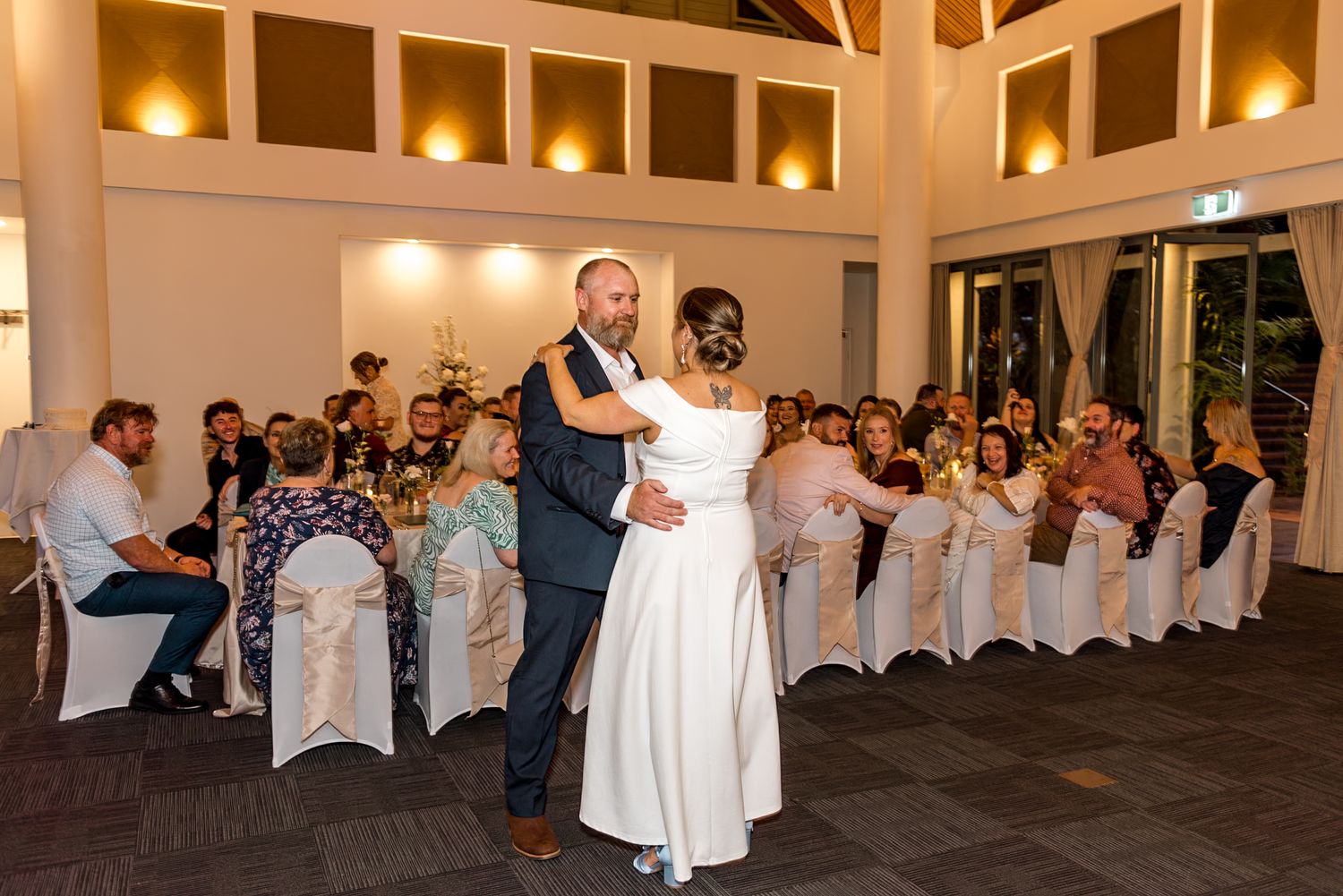 Newlyweds share romantic first dance in warmly lit wedding reception hall surrounded by seated guests.