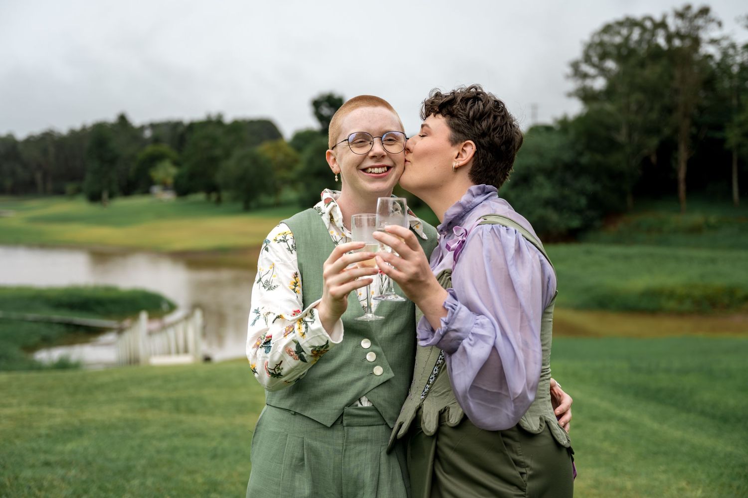 Two brides laughing and sharing a kiss while holding champagne glasses by a pond.