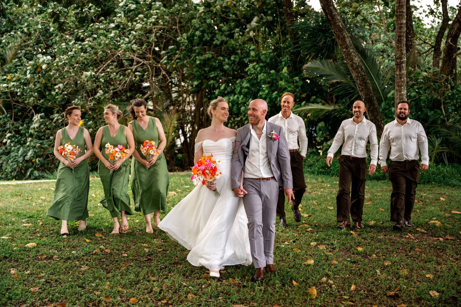 Wedding party strolls through tropical garden, bridesmaids in sage green gowns carrying colorful bouquets alongside bride and groom.