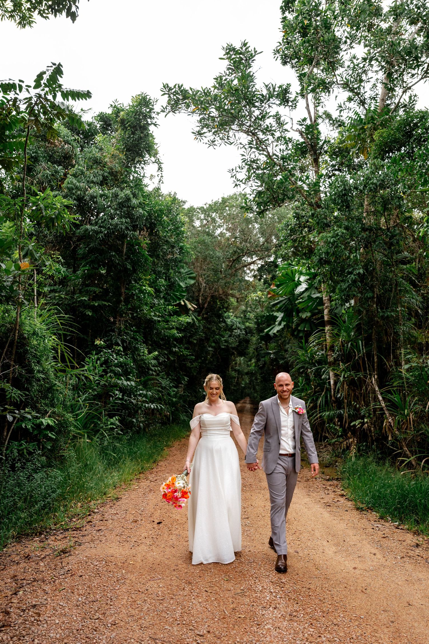Smiling bride and groom walking hand in hand down a dirt path framed by tall tropical green trees.