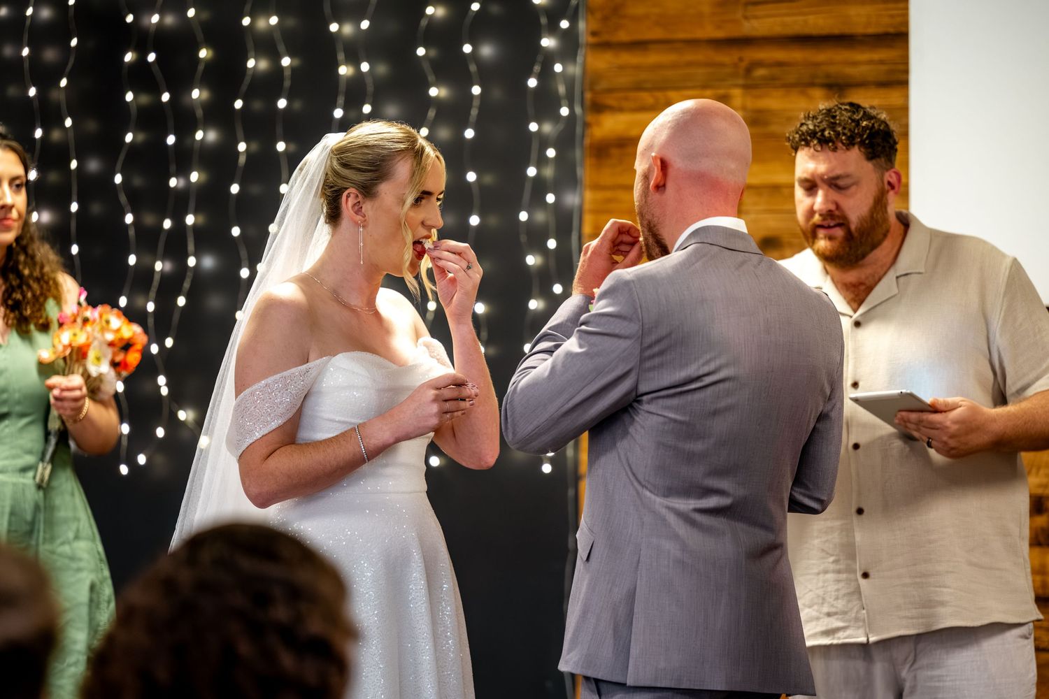 Bride and groom exchanging rings during wedding ceremony with officiant and fairy lights behind.