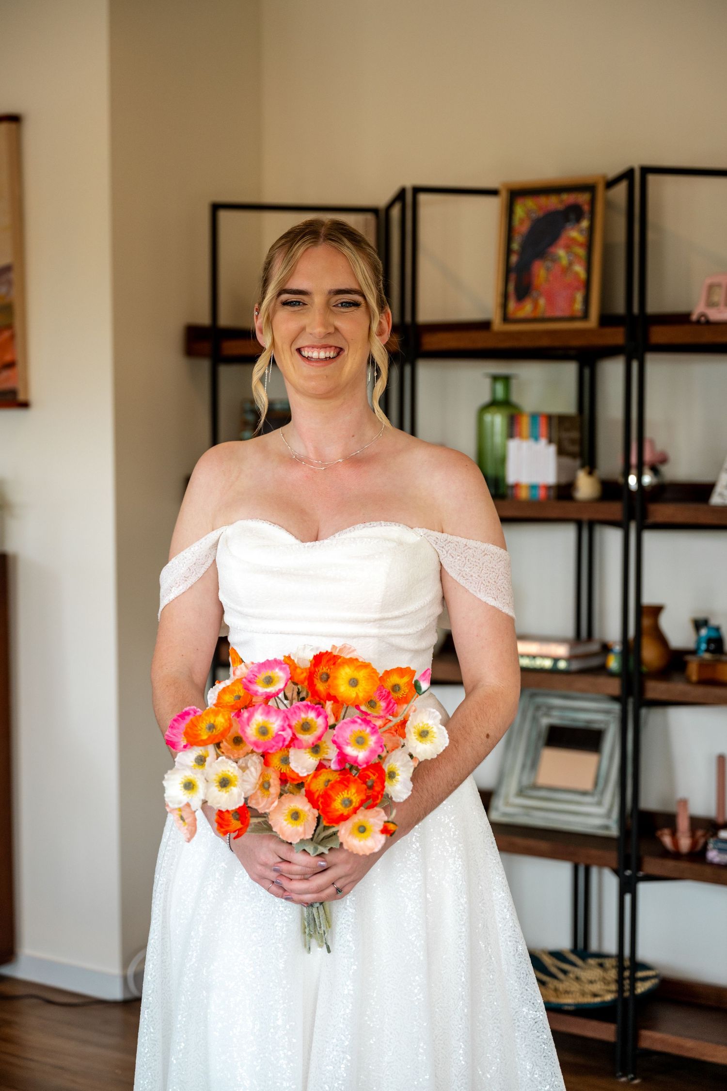 Smiling bride in white off-shoulder wedding gown holds vibrant orange and pink floral bouquet indoors.