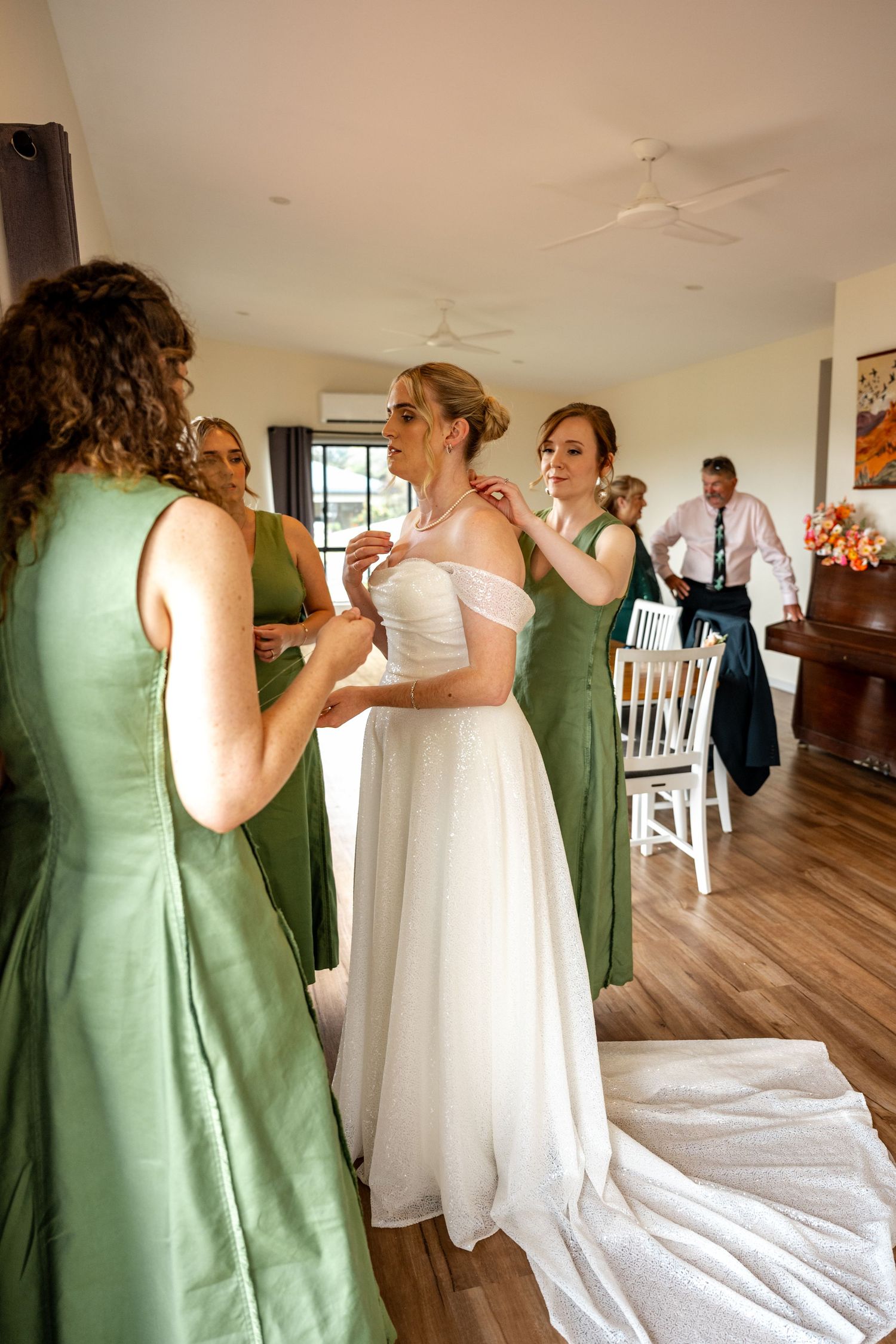 Bridesmaids in green dresses help bride with jewelry during wedding morning preparations.
