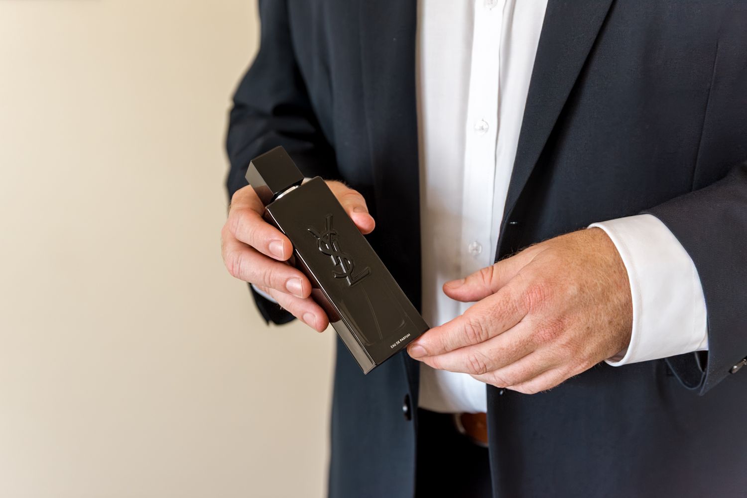 Close-up of groom in navy suit holding black cologne bottle with both hands.