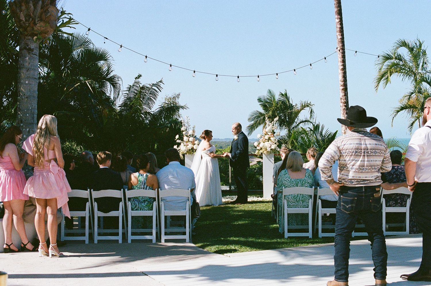 Outdoor beach wedding ceremony with guests seated in white chairs under string lights surrounded by palm trees.
