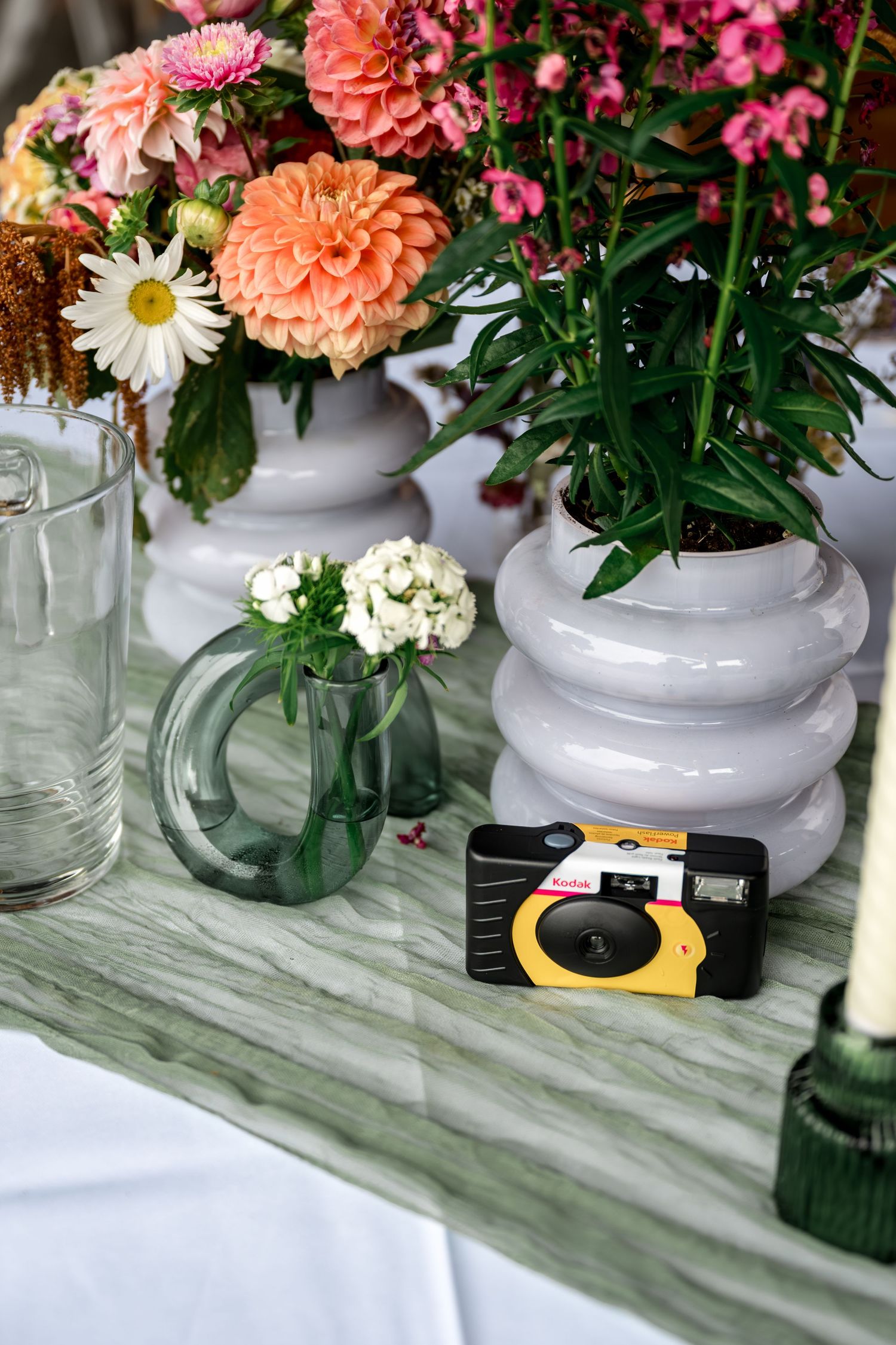 Wedding table display with dahlias, disposable Kodak camera, green vase, and white ceramic vessels.