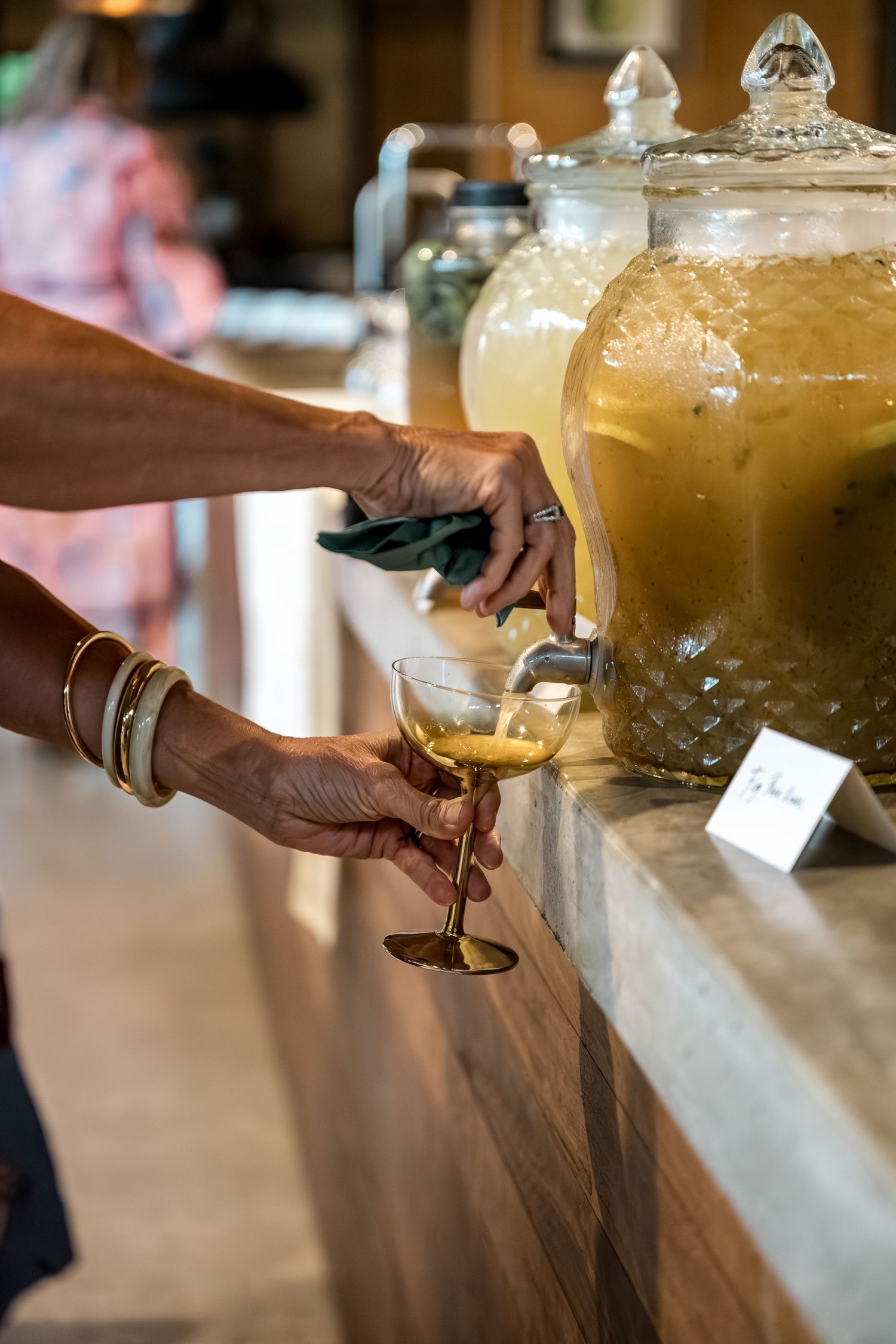 Woman pouring golden drink from large glass dispenser into coupe glass at wedding bar.