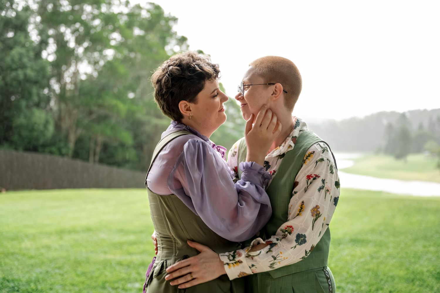 Two people in green outfits gazing at each other tenderly while embracing on a lawn.