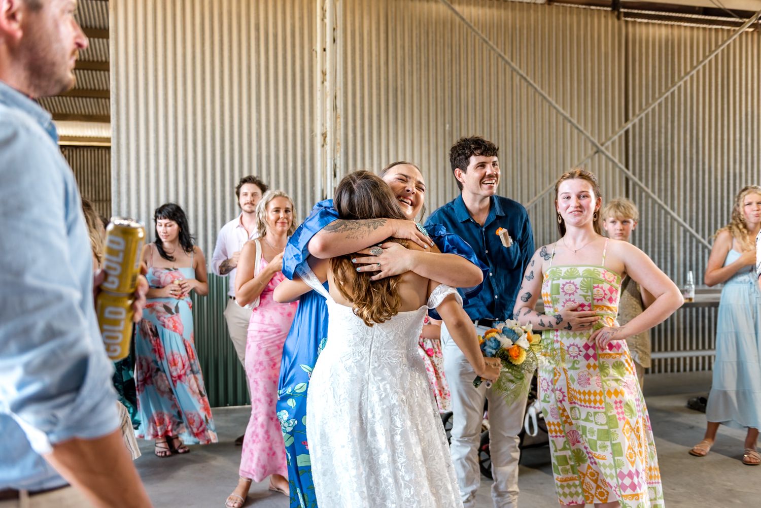 Bride in white lace gown receives warm hugs from excited wedding guests inside a rustic barn.
