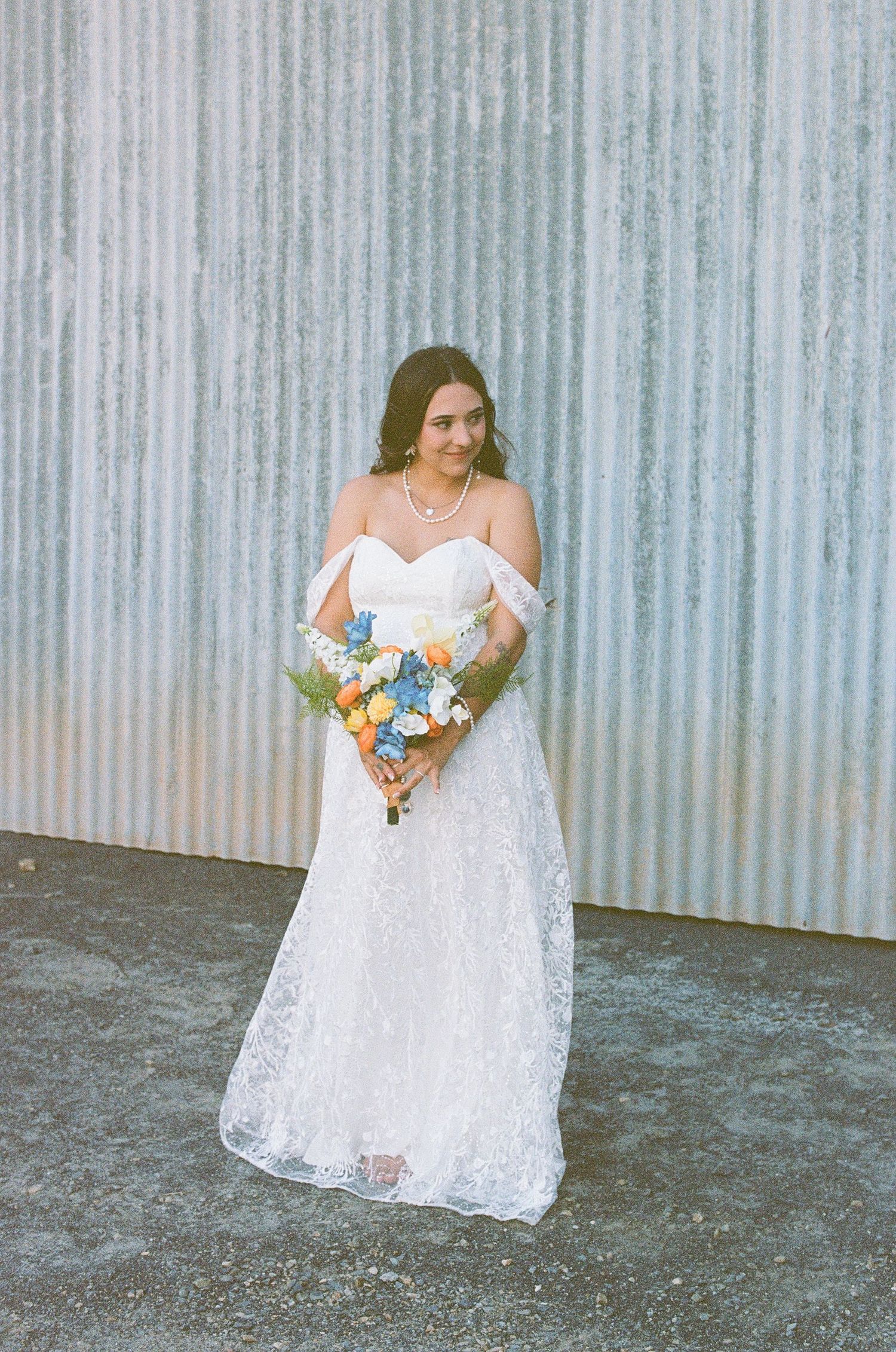 A bride in an off-shoulder lace wedding gown holds a colorful bouquet of blue and orange flowers by a corrugated wall.