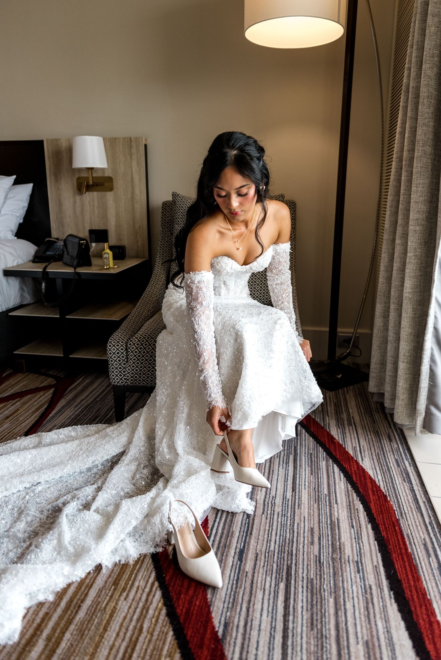 Bride in strapless lace wedding gown sitting in chair putting on white heels.