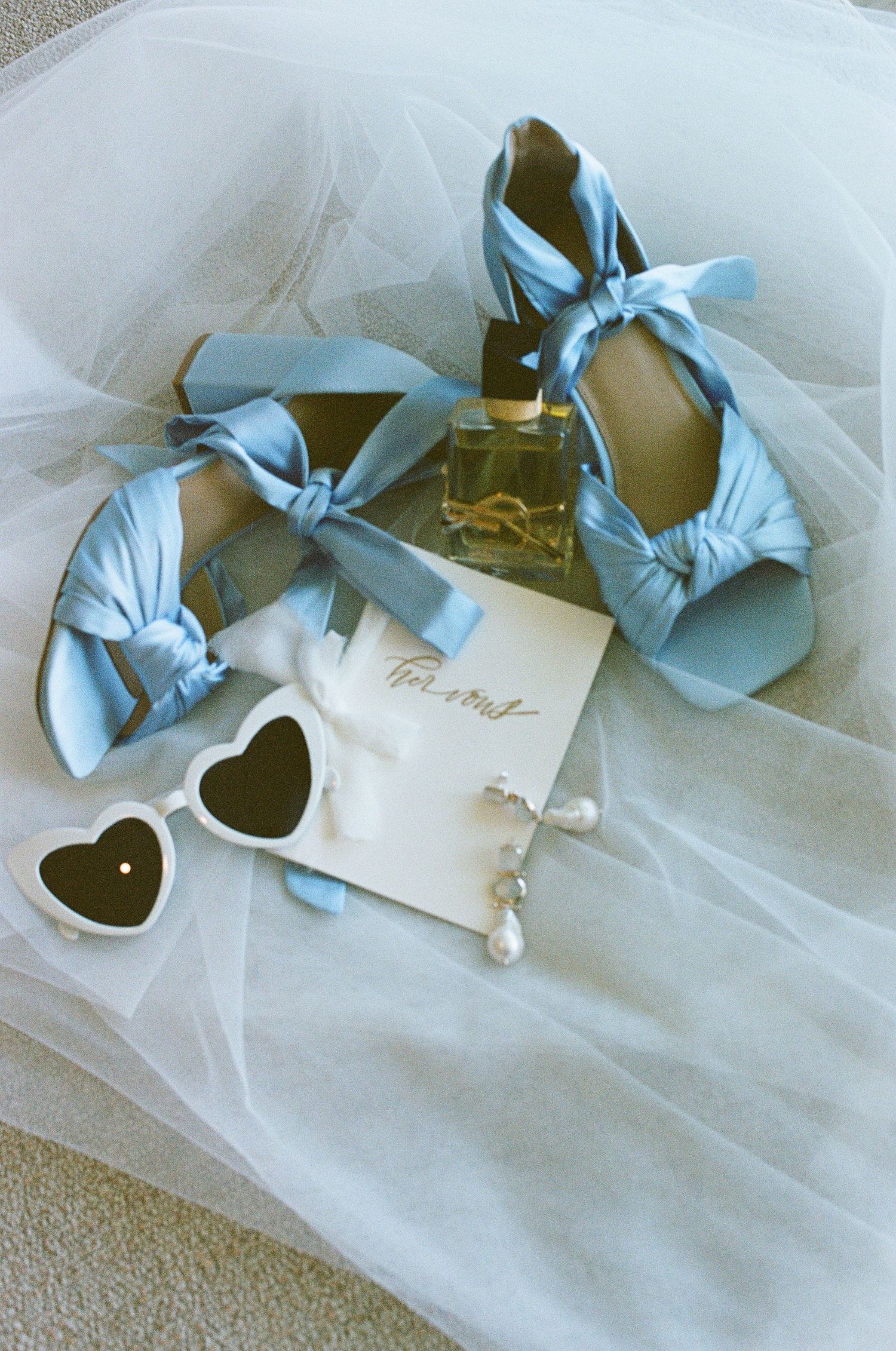 Bridal flat lay featuring blue satin bow heels, heart sunglasses, pearl earrings, and perfume.