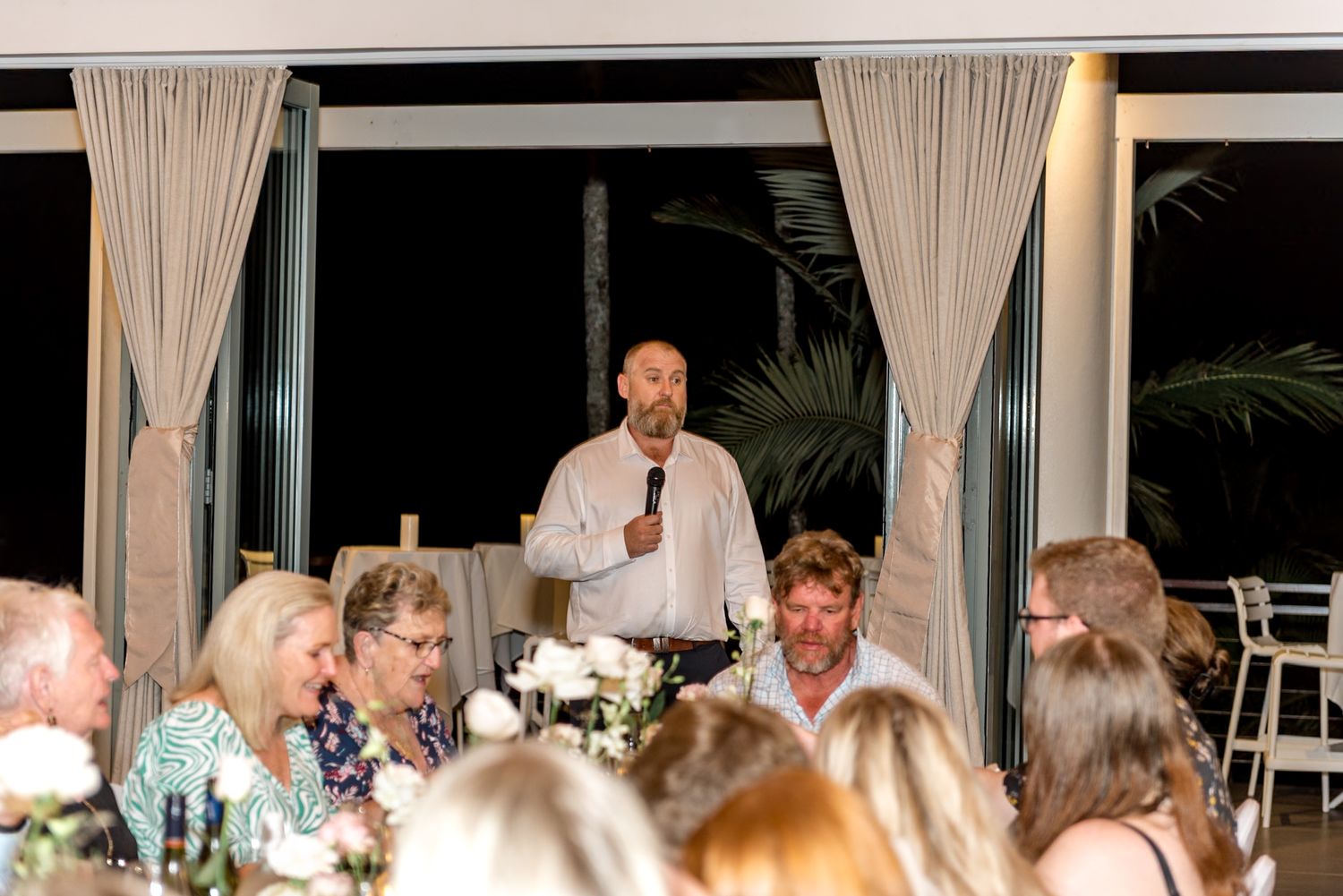 Man giving speech with microphone at wedding reception dinner surrounded by seated guests.