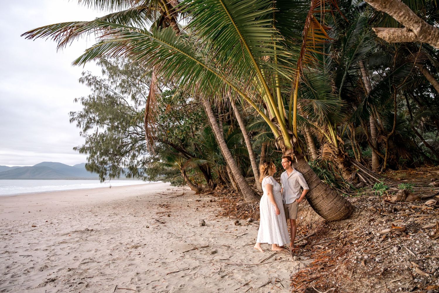 Couple in white outfits standing together on a tropical beach surrounded by palm trees at Four Mile Beach, Port Douglas.