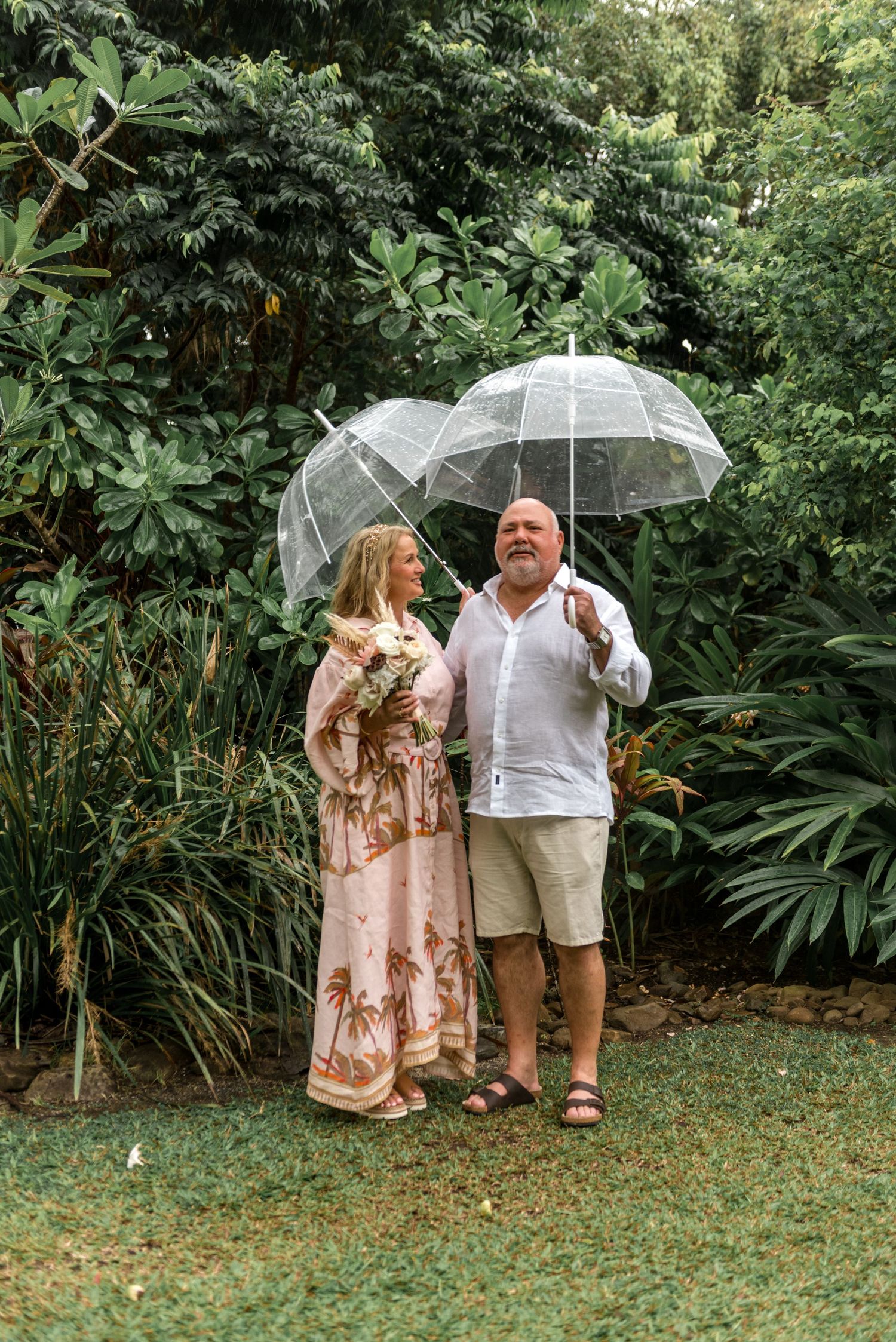 Mature couple standing under clear bubble umbrellas in a lush green tropical garden landscape.