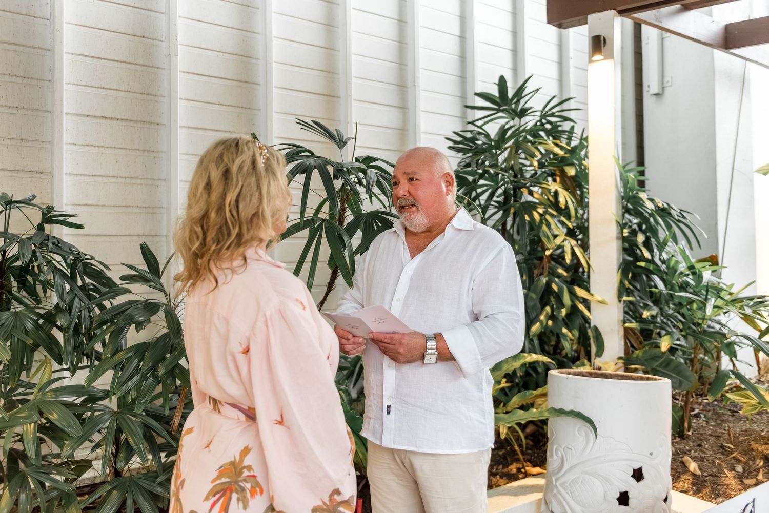 Man reads wedding vows to blonde woman in pink floral dress at outdoor ceremony.