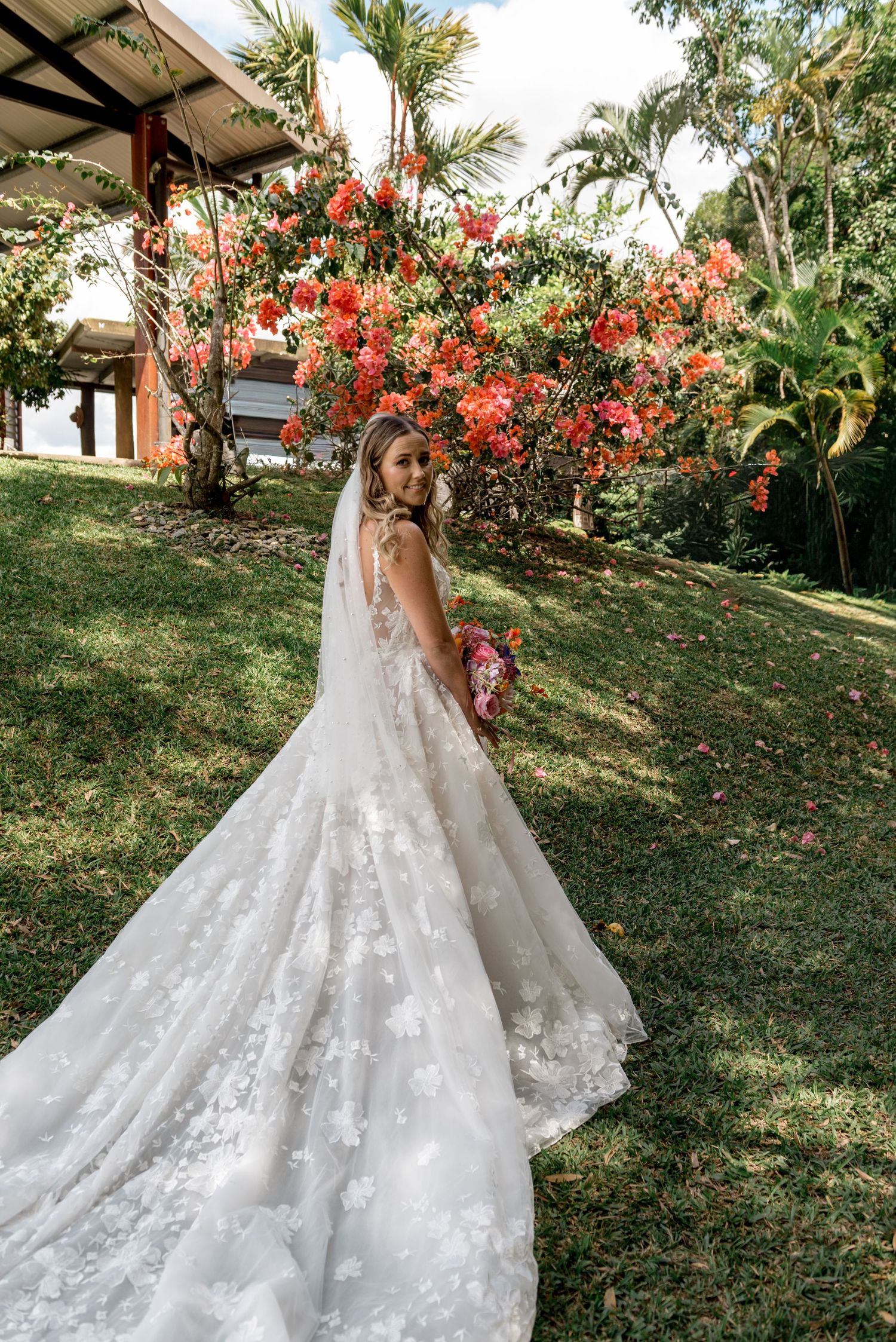Bride in floral lace wedding gown with cathedral train glances back near vibrant red flowering tree.