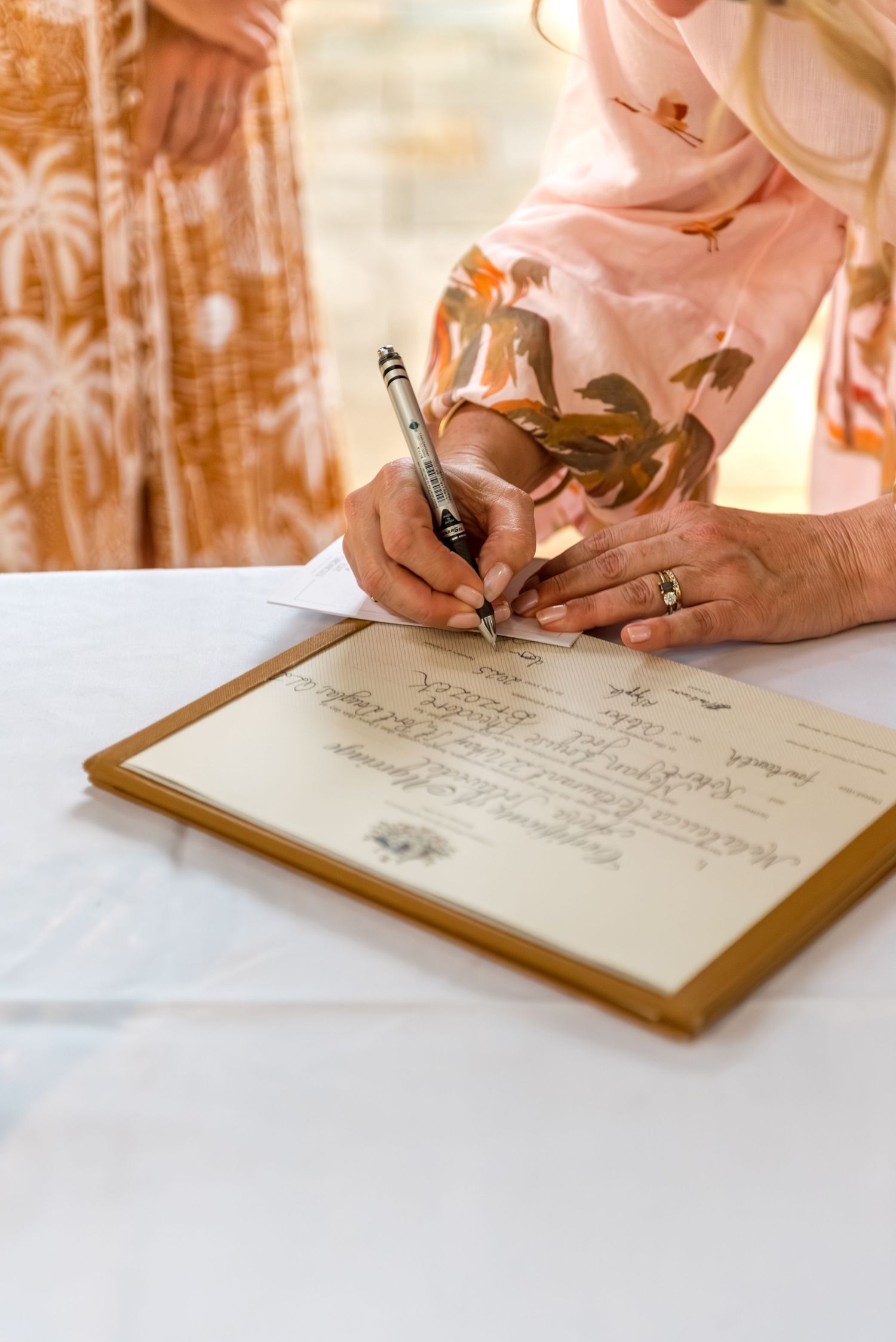 Close-up of bride signing wedding certificate with elegant pen, wearing diamond ring at white draped table.